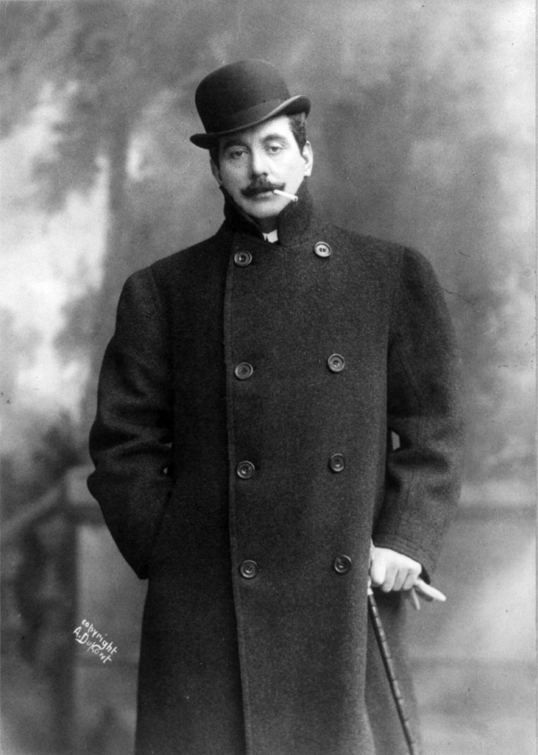 A greyscale image of Puccini wearing a bowler hat and overcoat, leaning on a cane, with a cigarette dangling from his mouth. 