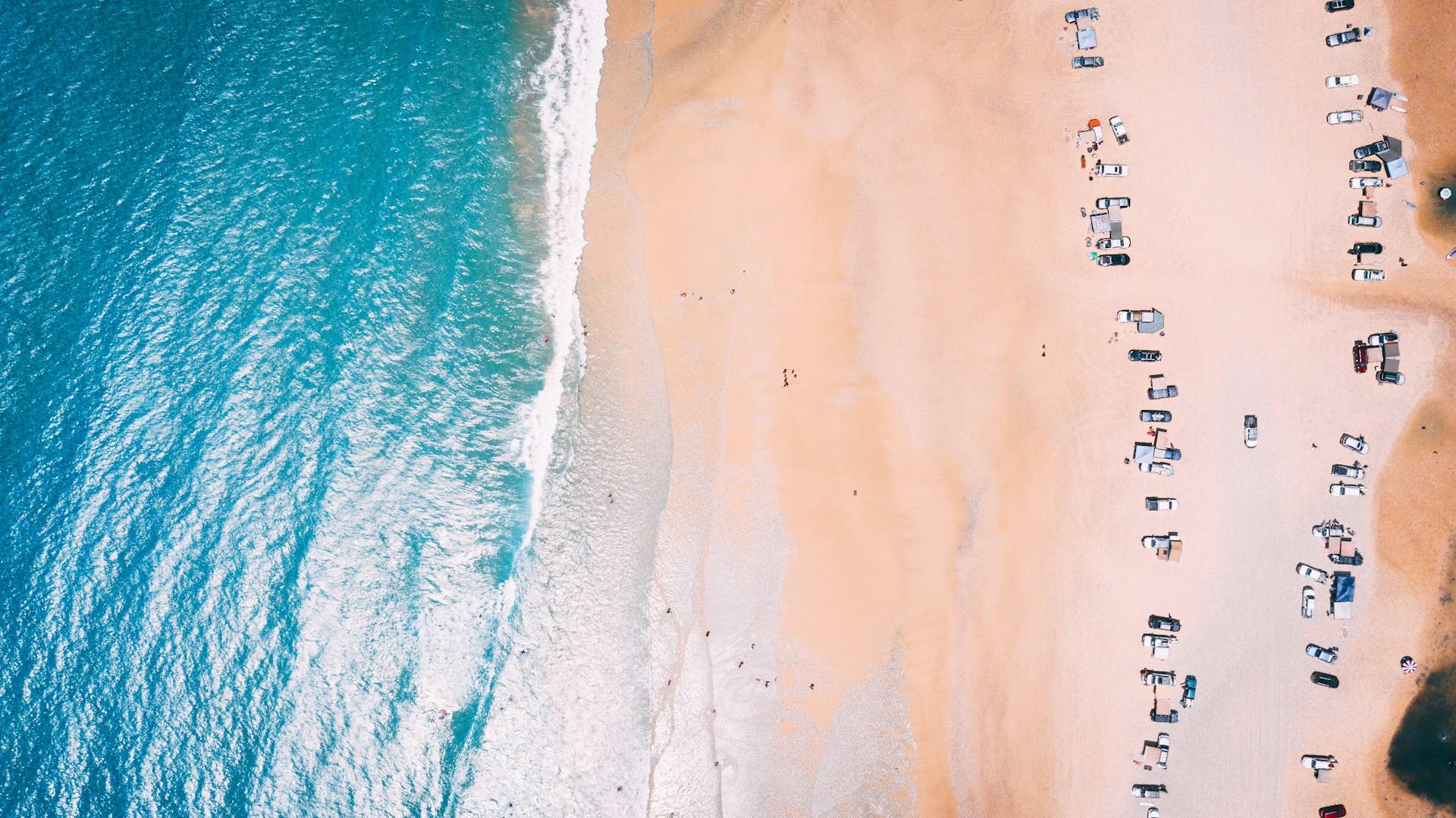 An aerial view of Brisbane's Rainbow Beach on a sunny day, with the sand appearing orange in contrast with the blue water and white froth, and a row of beachgoers along the tide line.