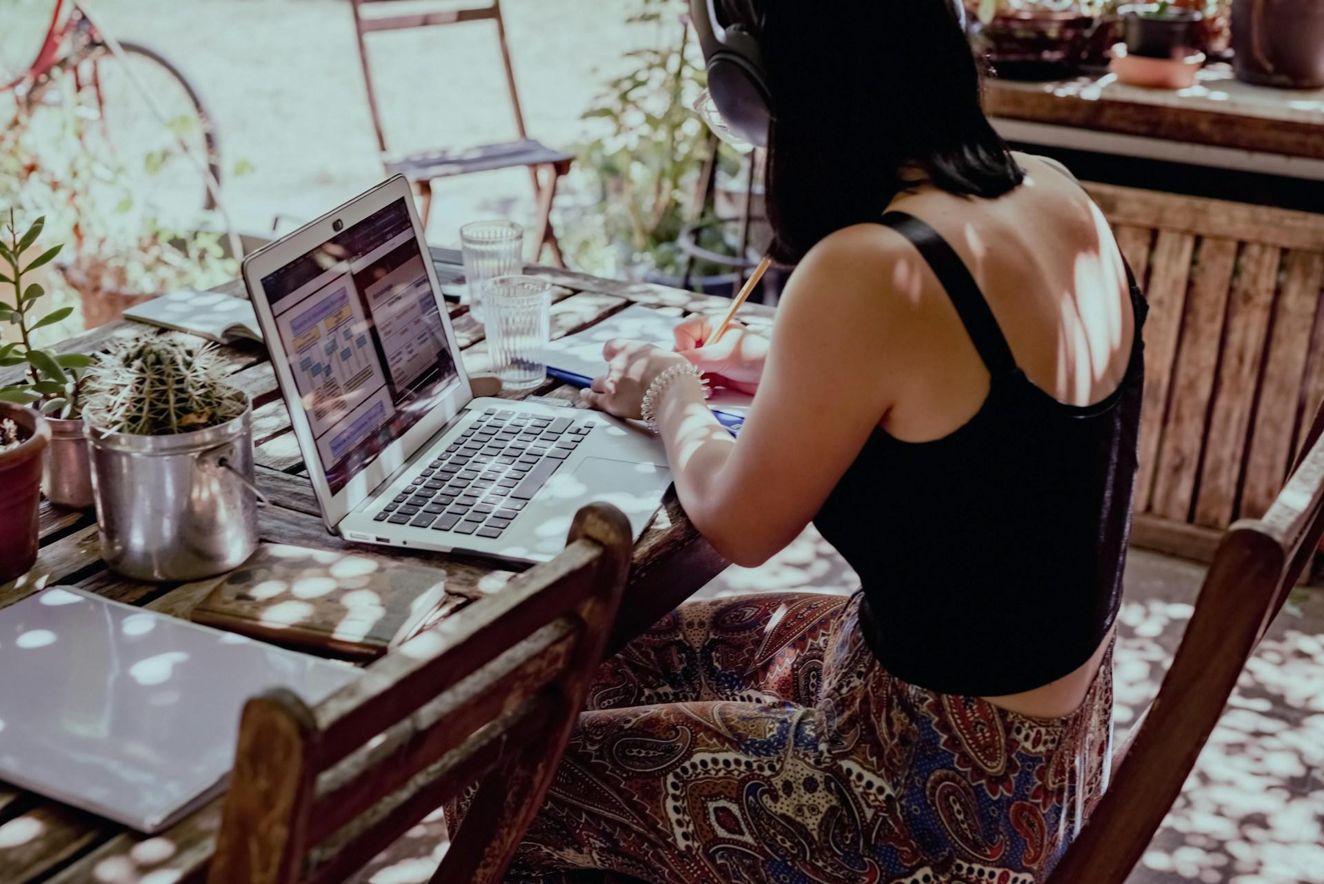 A person wearing a black top and paisley print bottoms sits at a wooden table dappled by sunlight, with an open laptop computer in front of them, writing with the yellow pen they hold in their right hand.