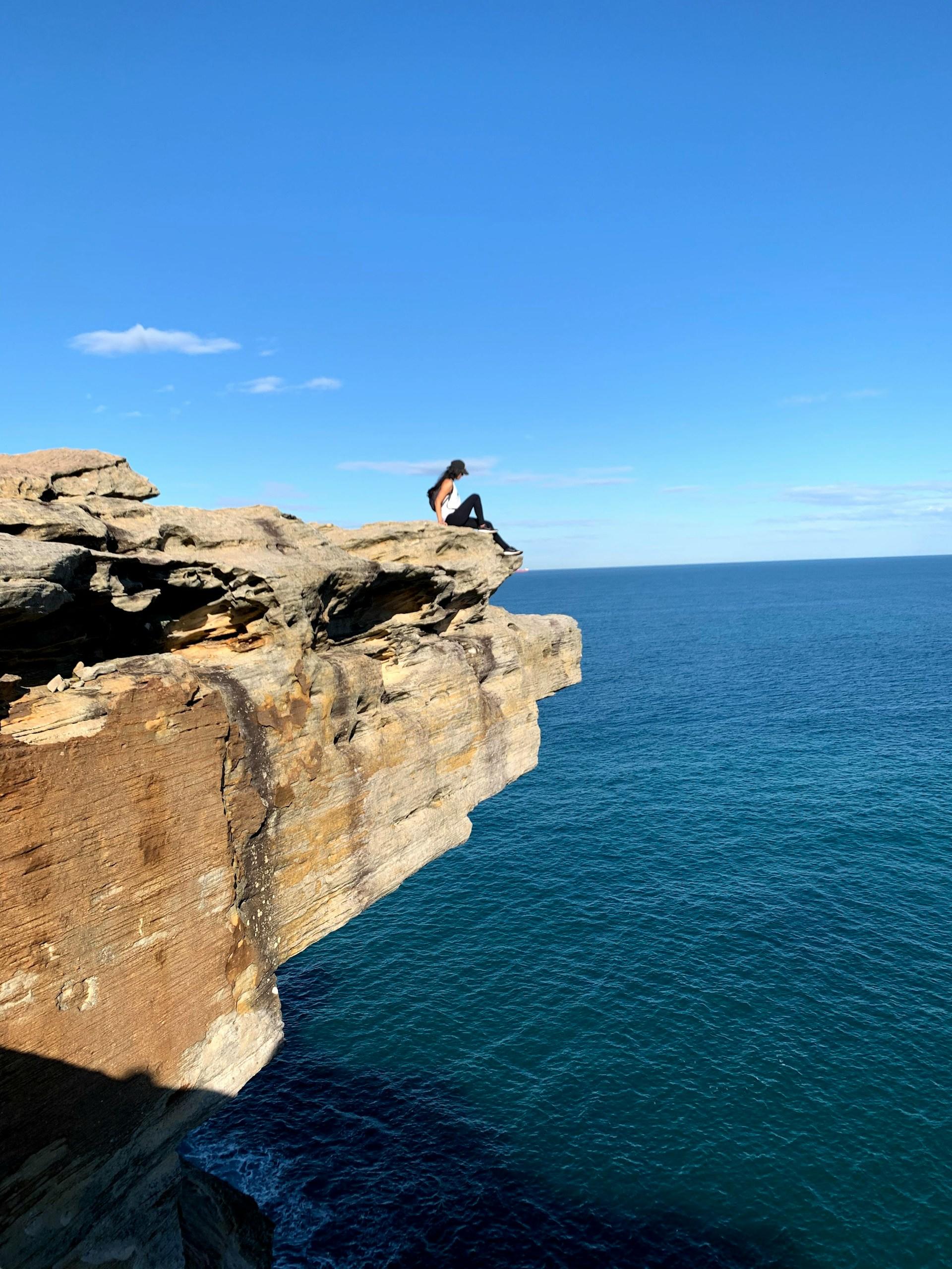 A person perched at the very tip of a sandstone outcrop that hangs over a deep blue body of water in Royal National Park, NSW.