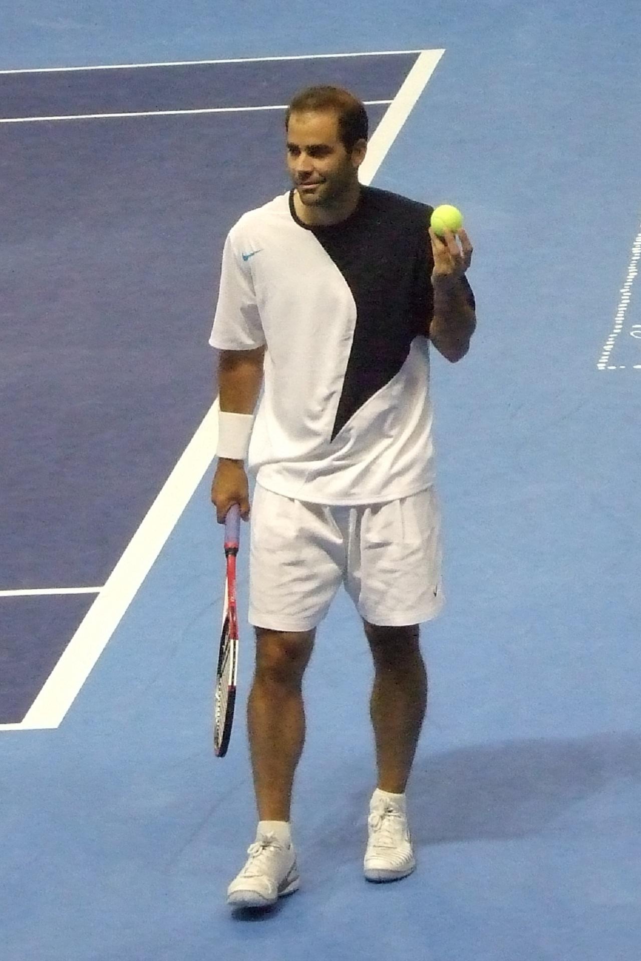 A man wearing white shorts with a white and black shirt holds a tennis ball and a racket.