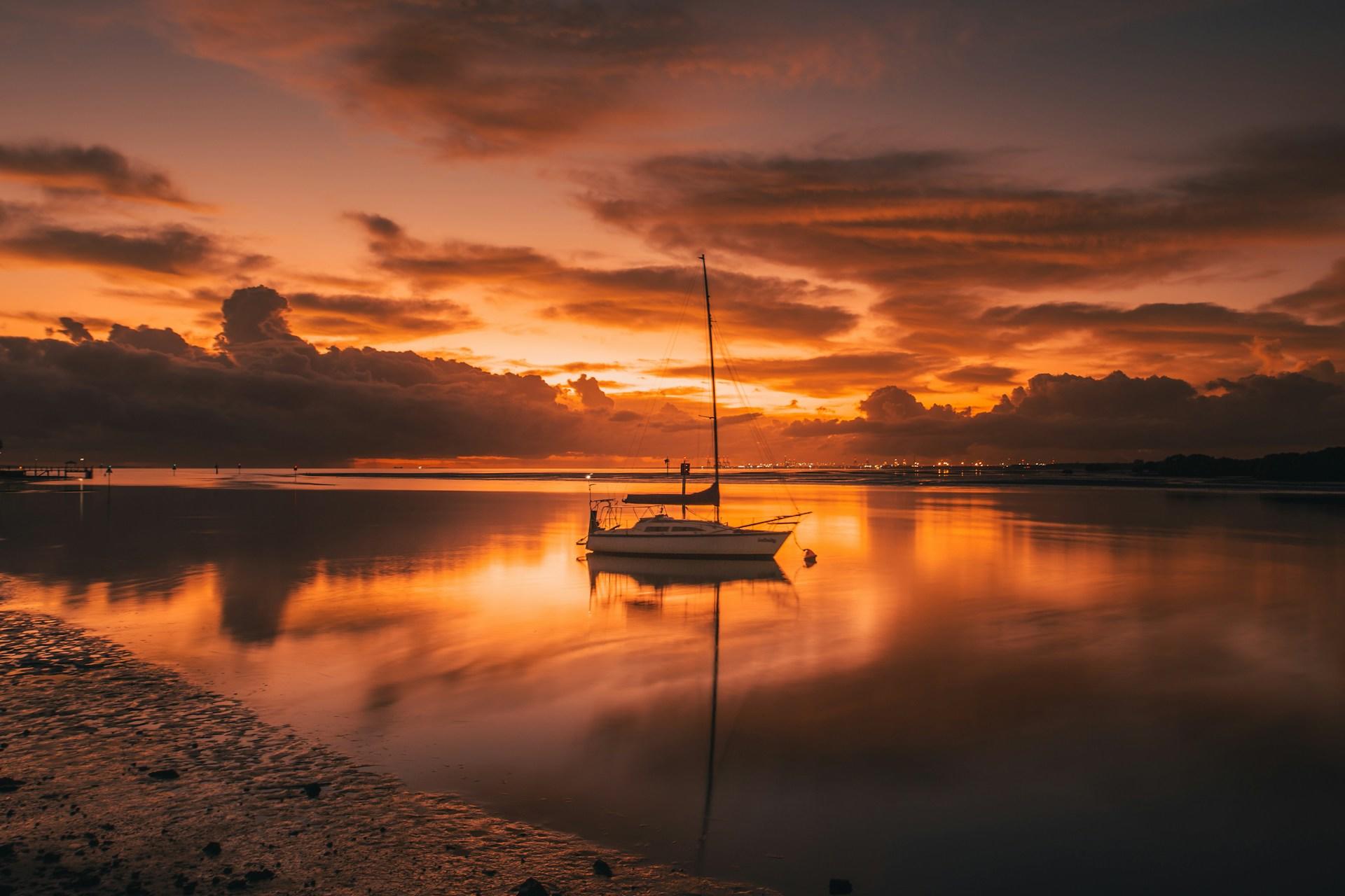 The Shorncliffe strand and still waters under a deep orange sunset, with a lone sailboat, sails furled, at anchor near the shore.