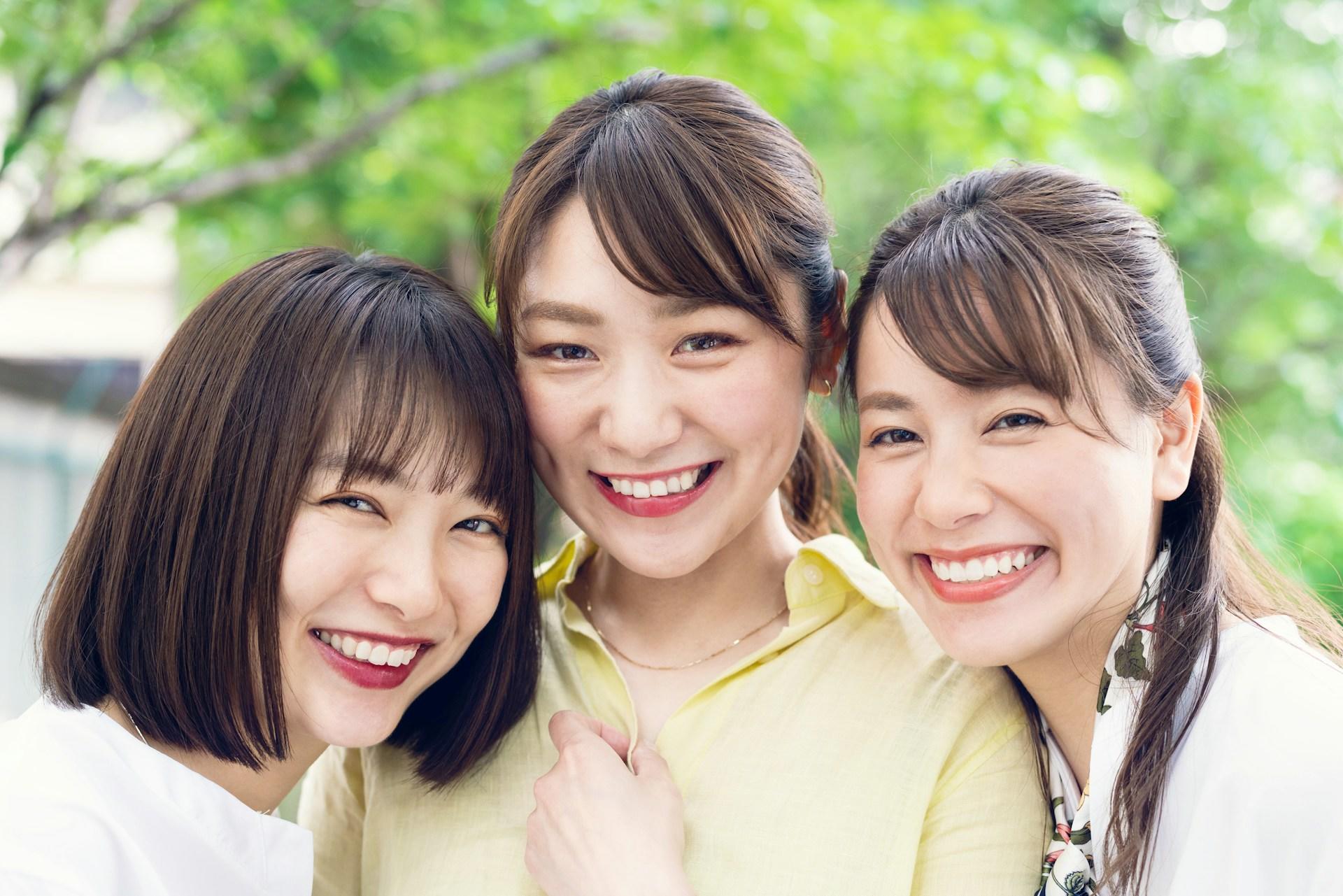 Three smiling women huddle together to face the camera. Two have their long hair pulled back while the third wears a pageboy cut. All three wear light-coloured clothes against a leafy green background