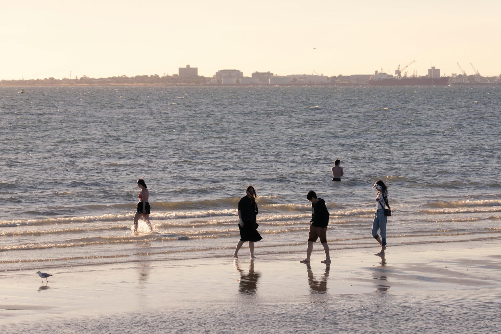St Kilda Beach at sunset, with relatively calm waters and a few beachgoers wearing street clothes strolling along the sand and wading in the shallow water at sunset.