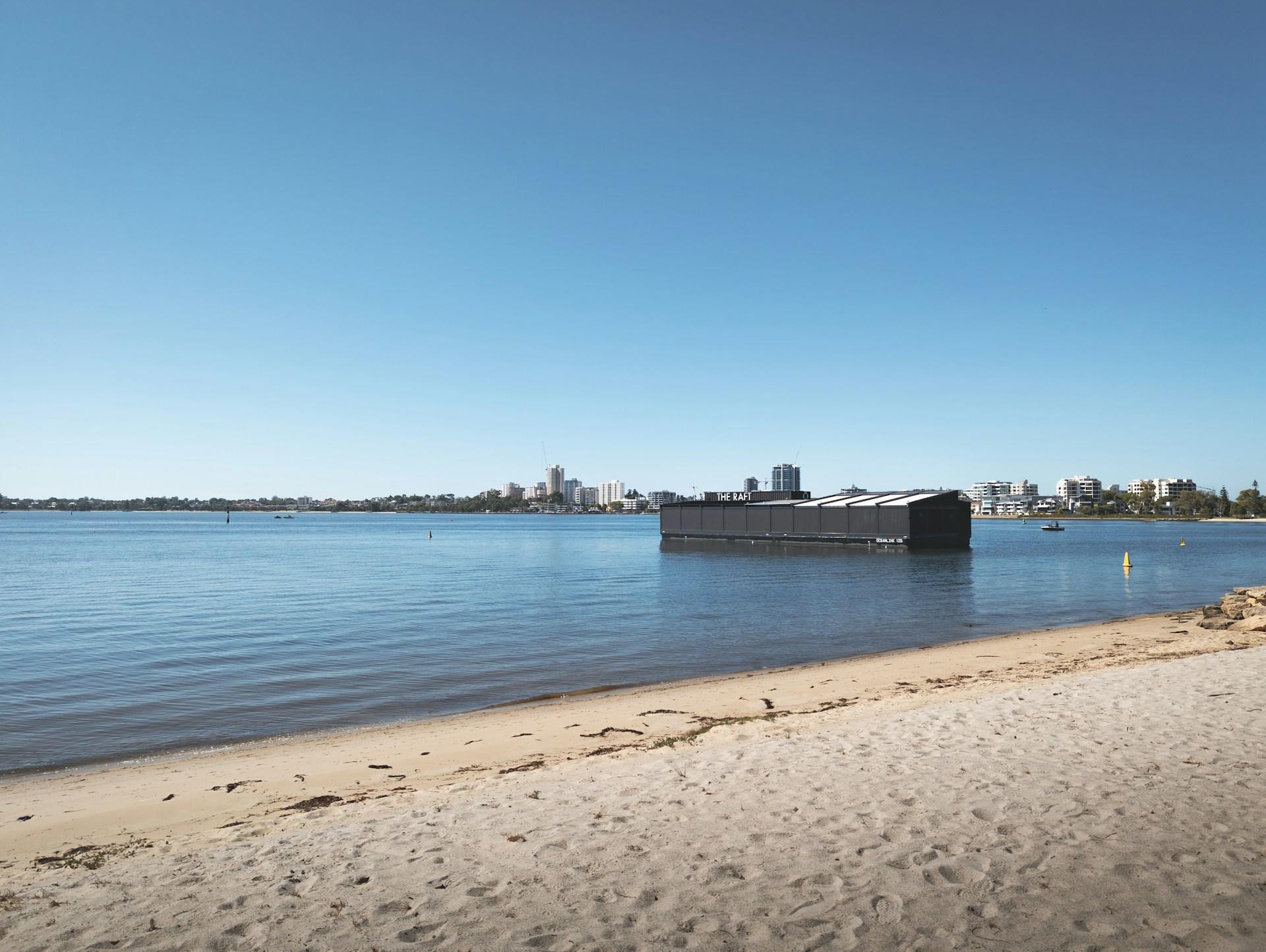 A view of The Raft, a black structure a few metres off the Swan River beach, seen in daylight from the deserted beach's perspective, with white buildings rising up on a spit of land behind it. 