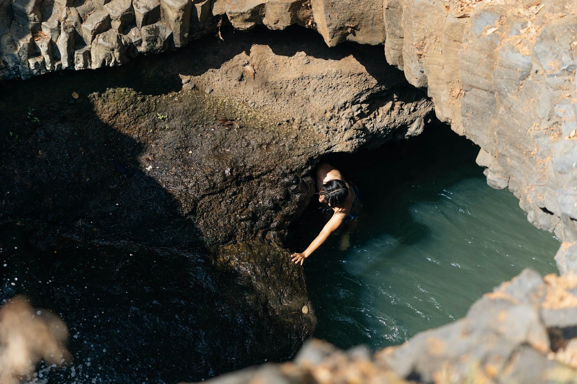 A person with bare arms stands in a shallow body of water just outside of a cliff fall, bracing themselves on a boulder in front of the cliff face on a sunny day.