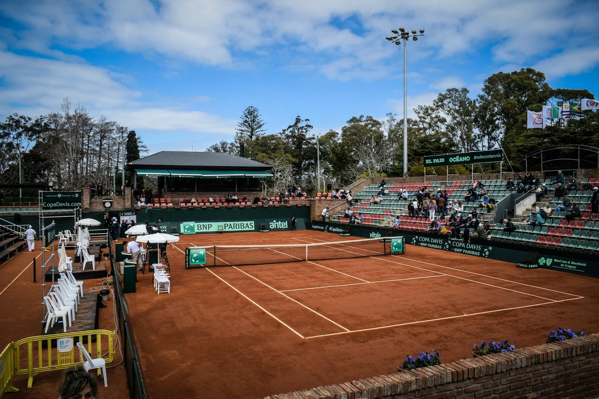 An open-air clay court with a few spectators in the green and red stands, while white chairs on the opposite side of the court stand empty and the court is devoid of players.  