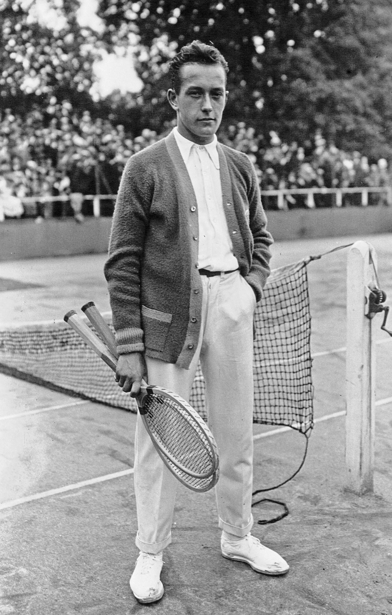 A greyscale image of a male tennis player holding two rackets and standing by the net. 