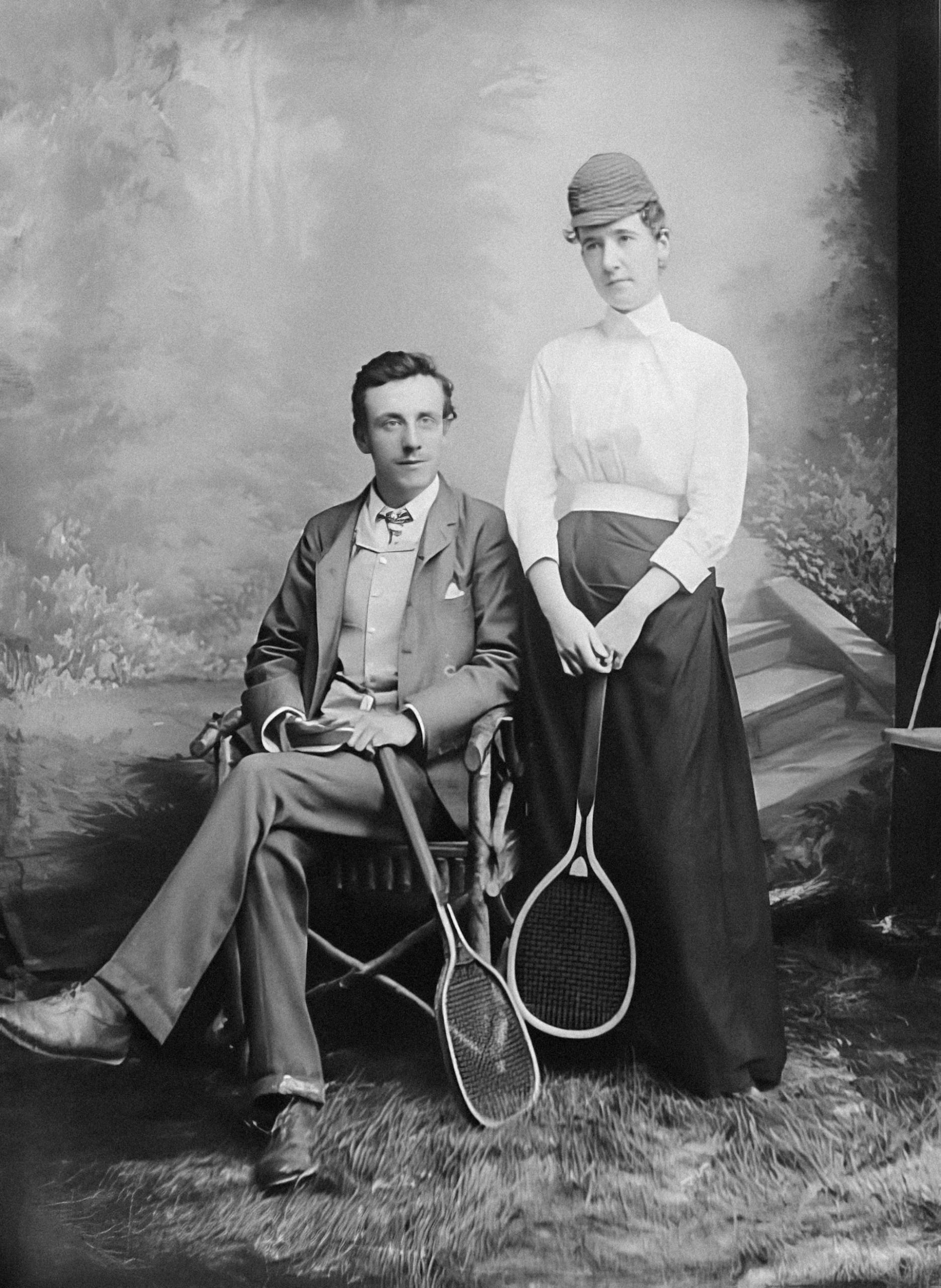 A greyscale image of a couple wearing tennis garb and holding rackets, with the man seated and the woman standing to his left.