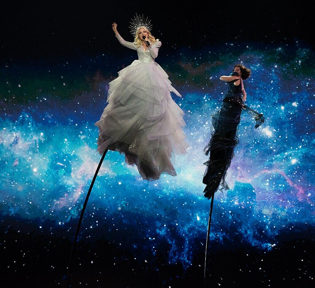singer Kate Miller-Heidke and backup singer supported on thin poles to appear as if they are floating in space against a galaxy background image.