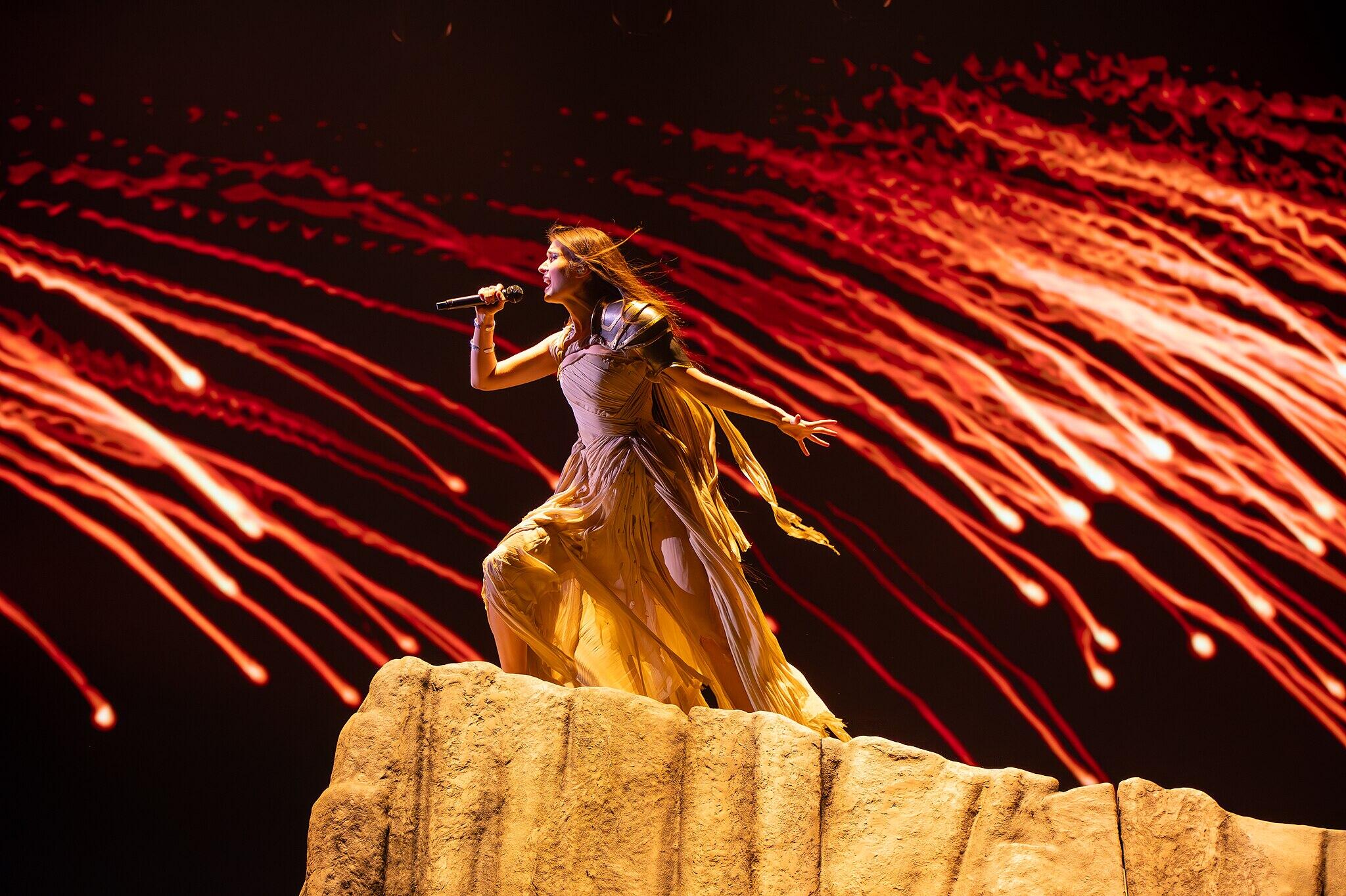 Singer Alyona Alyona standing atop a rocky stage prop with red fireworks in the background