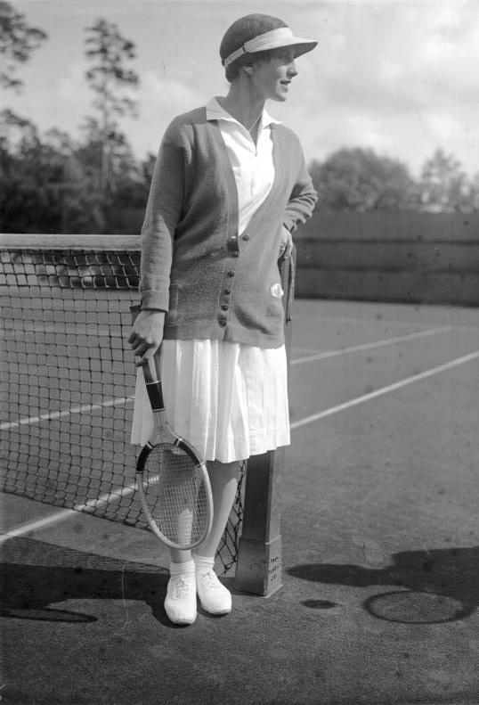 A greyscale image of a female tennis player in a knee-length skirt and cardigan on a tennis court.