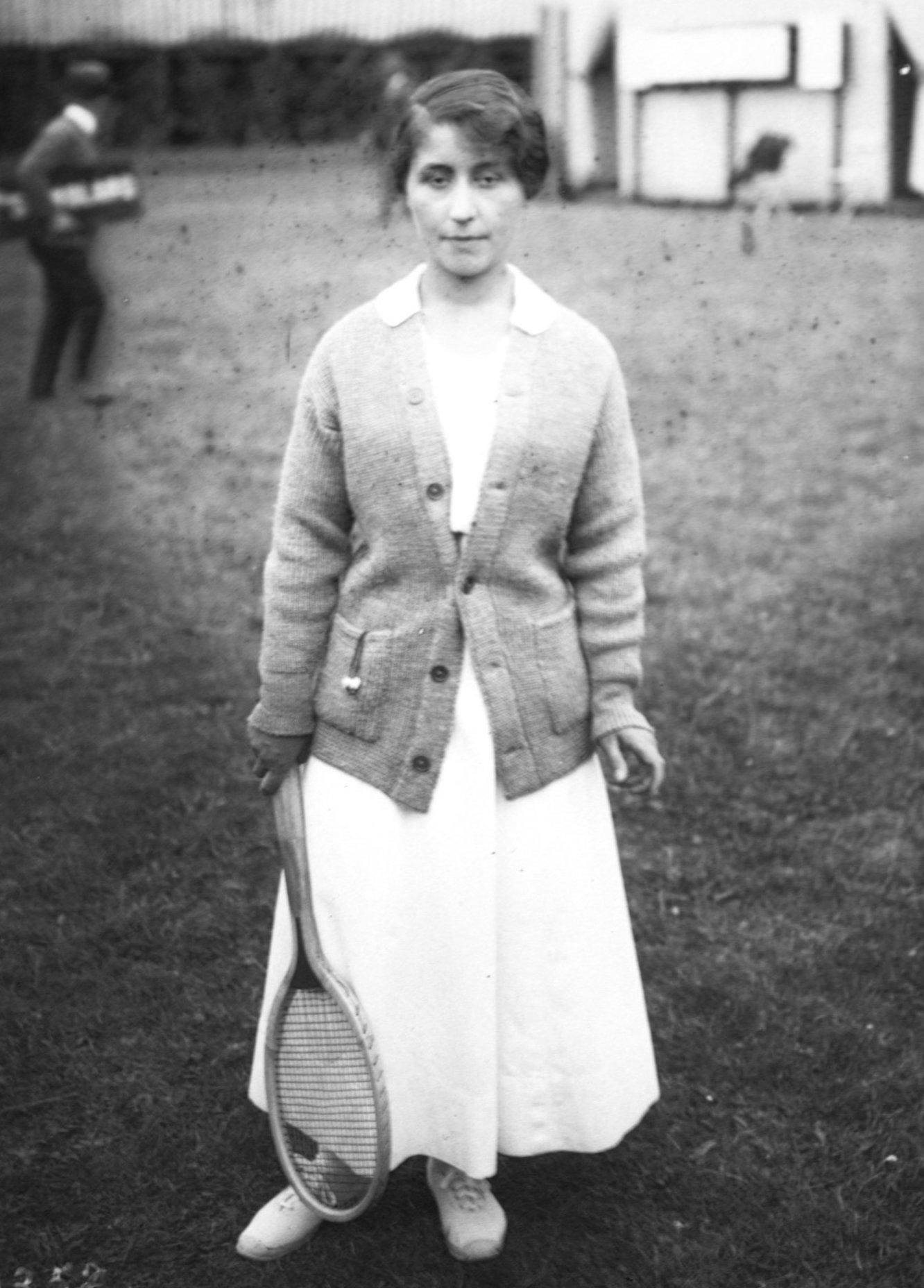 A greyscale image of a female tennis player in a long skirt and cardigan holding a tennis racket.