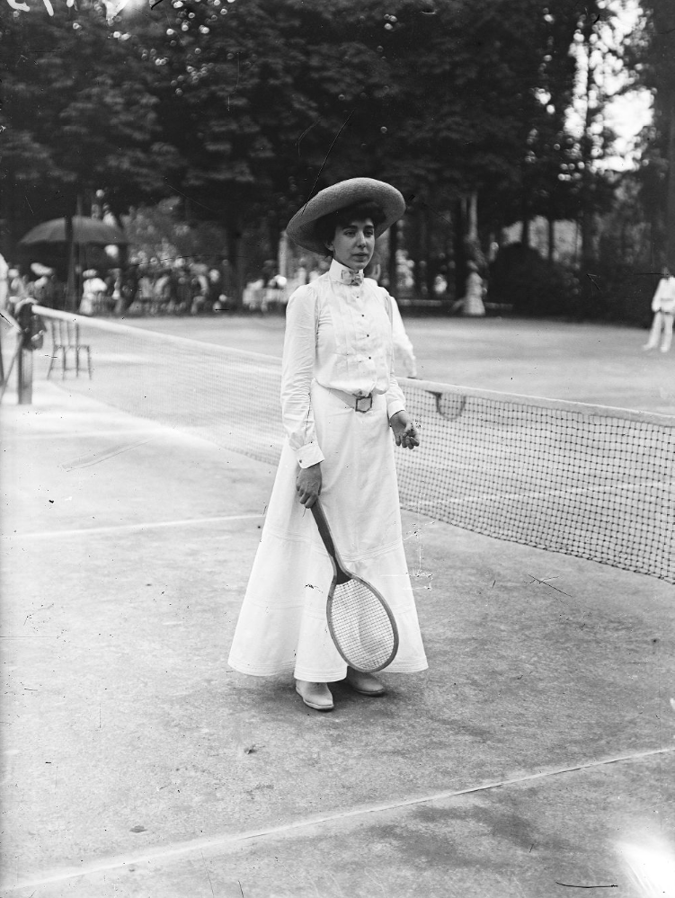 A greyscale image of a female tennis player wearing a hat and long skirt on a tennis court holding a racket.