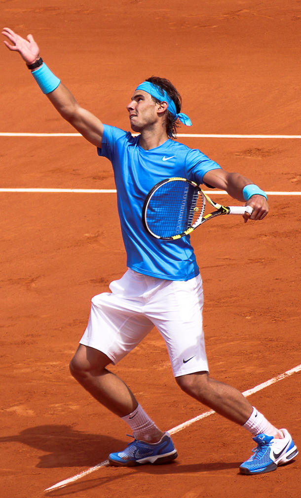 Rafael Nadal, wearing a blue jersey with white Nike swoosh on the left breast and white shorts, prepares to serve the ball on a clay court.