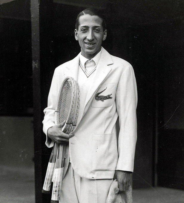 A greyscale image of a male tennis player wearing a blazer and holding two rackets.  