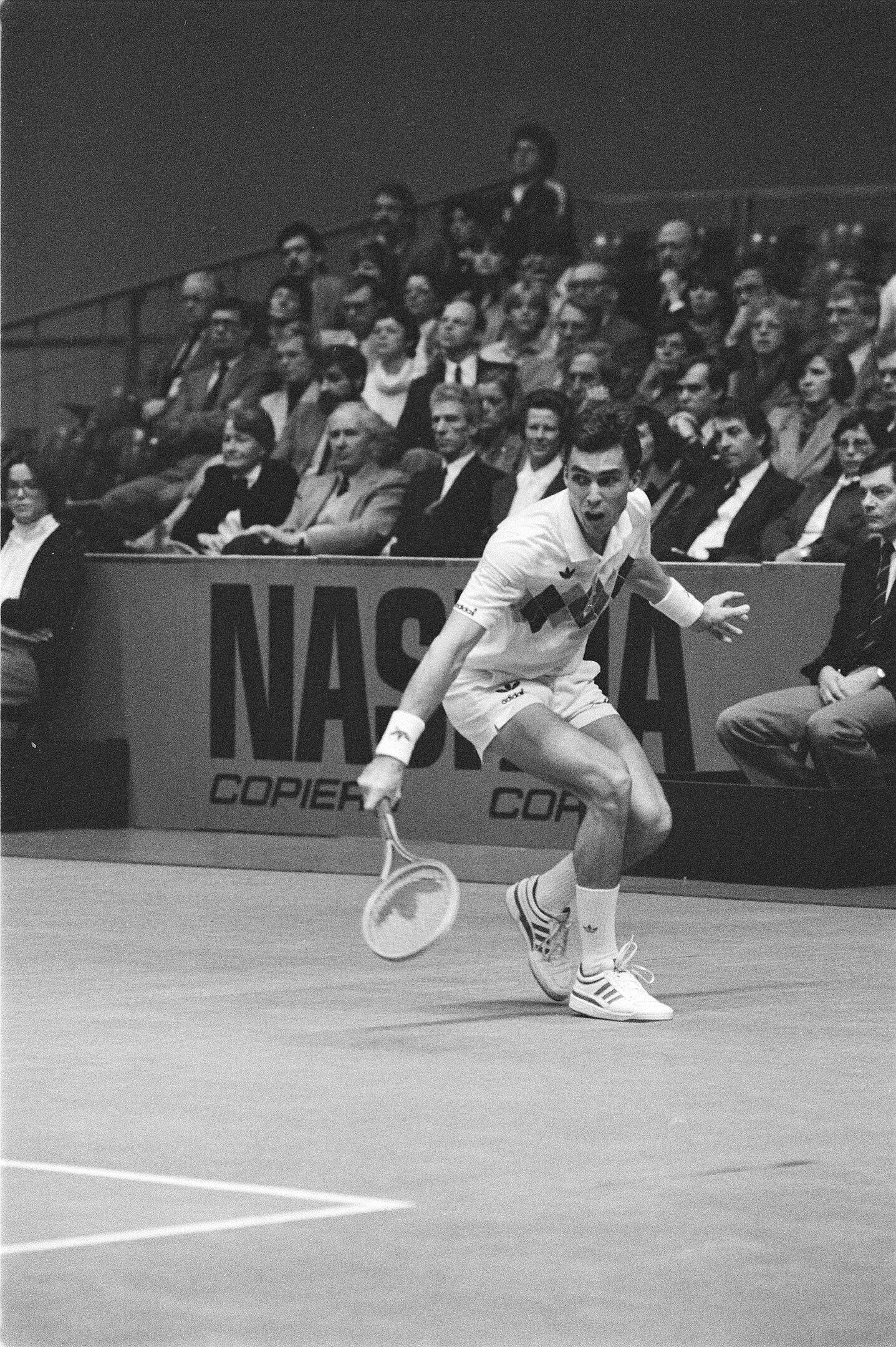 A male tennis player on an indoor court returning a ball, with spectators behind him. 