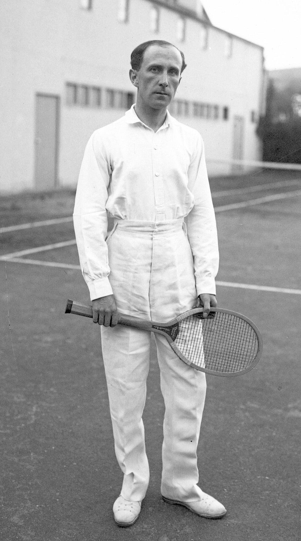 A male tennis player wearing whites and holding a racket on an outdoor court. 