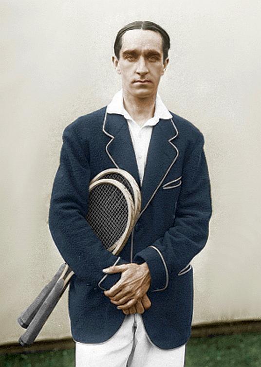 A male tennis player holding two rackets and wearing a blue blazer.