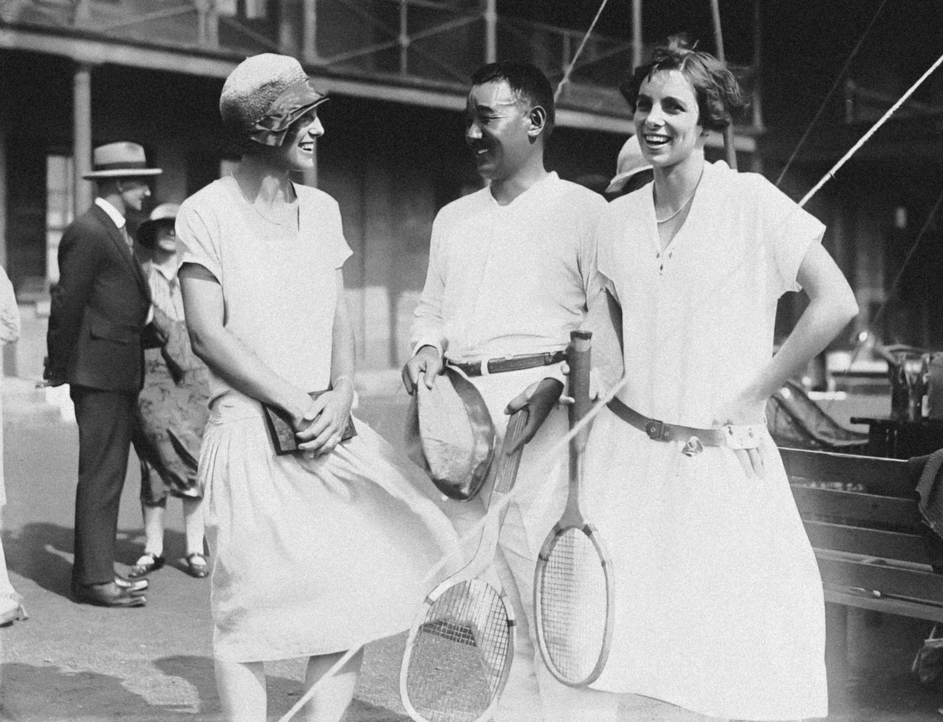 A greyscale image of three people dressed in tennis whites, two of them holding rackets and smiling. In the background, a man in a suit and hat stands in front of a building, next to a woman wearing a dress and hat. 