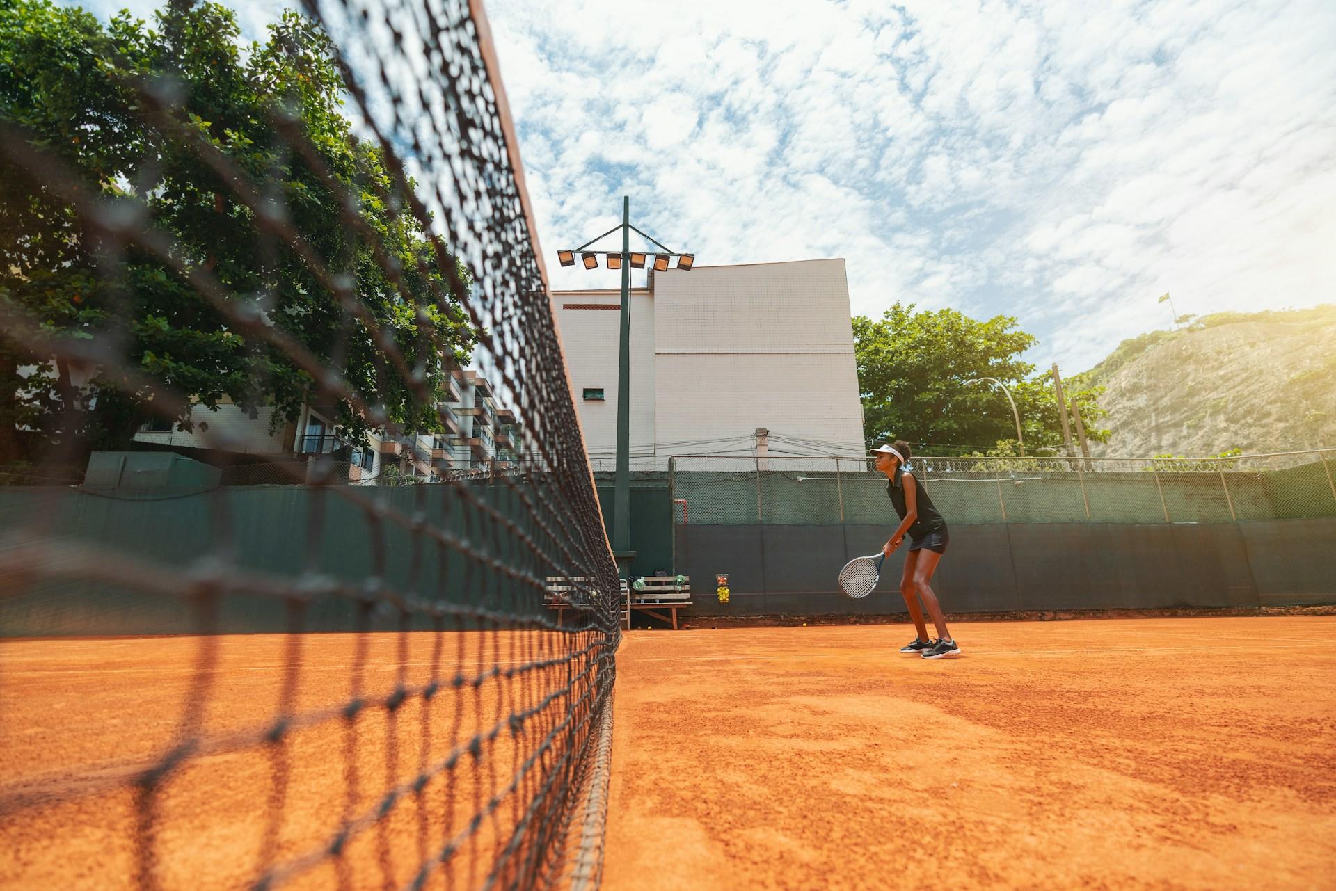 A long shot along a black net bisecting a clay tennis court, with a player wearing black standing at the far end and the white facade of a building, ringed by trees, in the background.