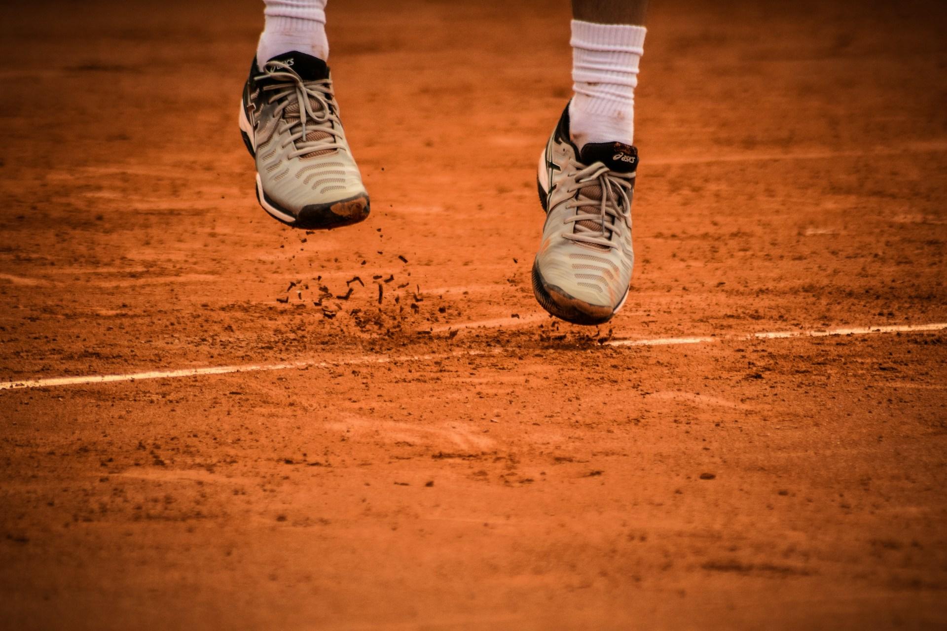 A tennis player's feet lift off the clay court as they jump to serve the ball.