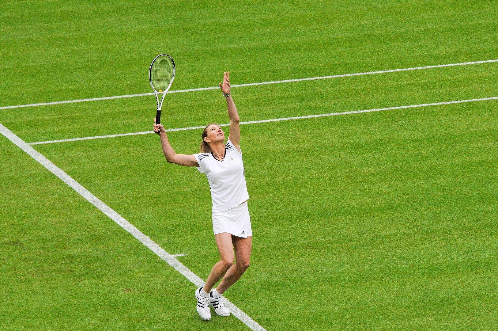 A female tennis player, wearing tennis whites, serving on a lawn tennis court.