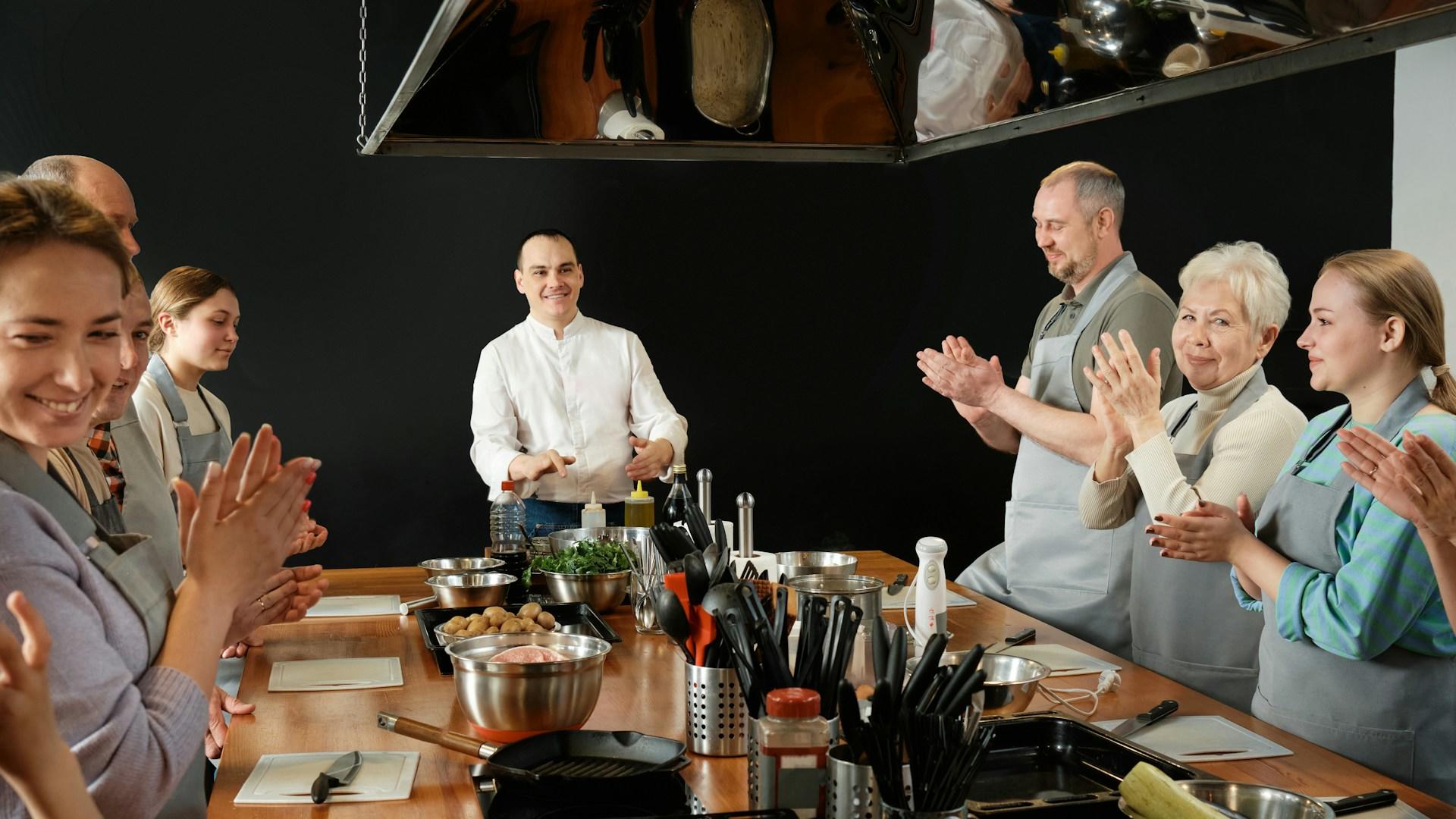 A group of people stand around a wooden table laden with food preparation equipment.