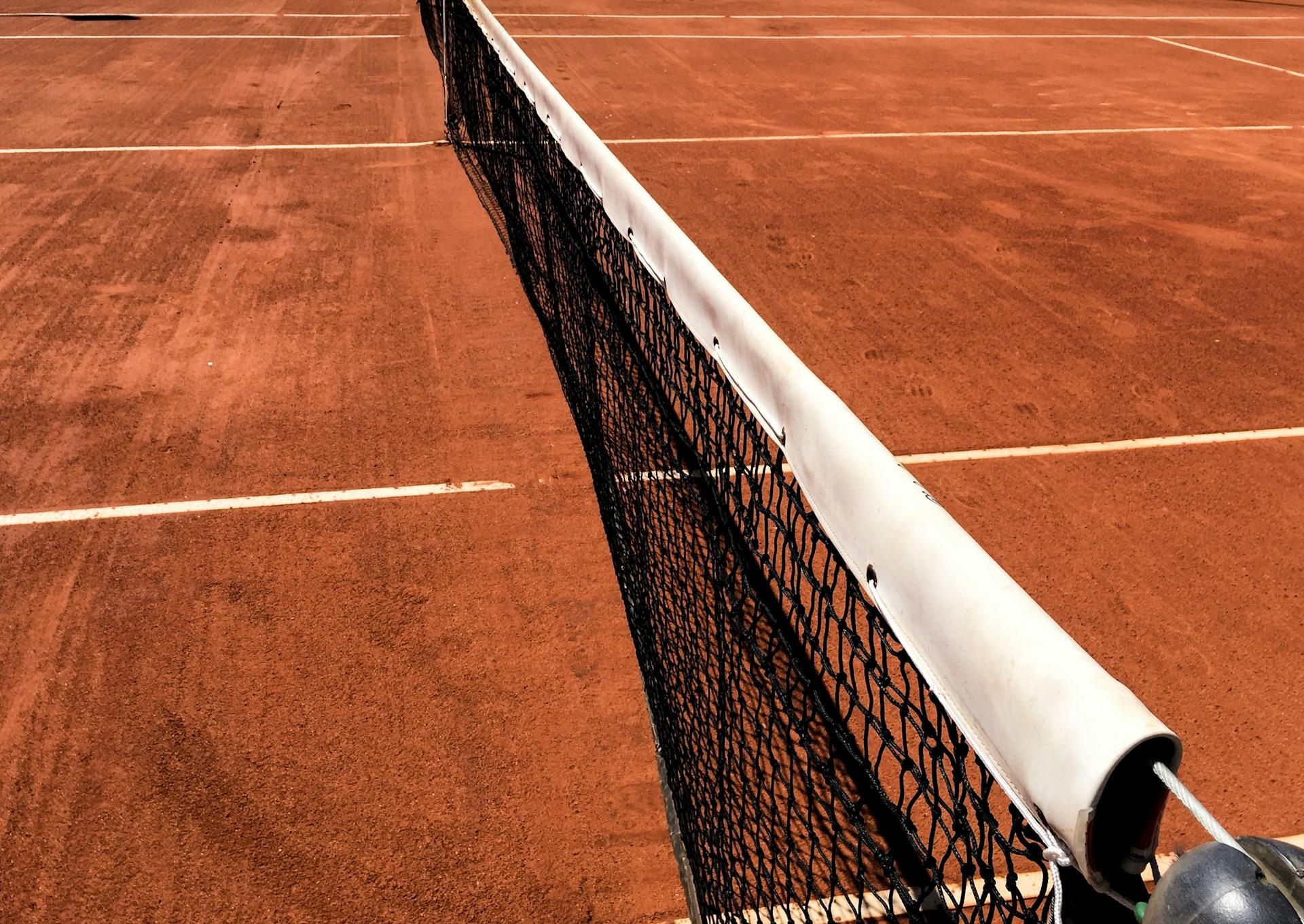 A close-up view of a black tennis net with a white top band bisecting a clay tennis court.