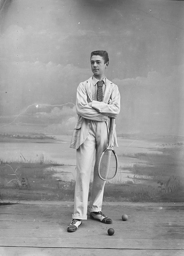 A person holding a tennis racket and a suit and tie posed in front of a beach scene background. 