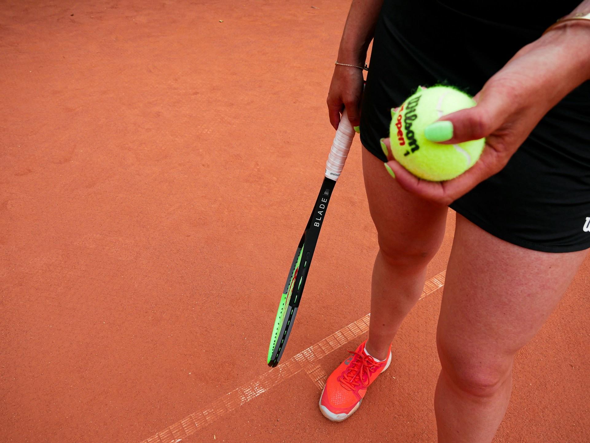 A tennis player wearing orange shoes and a black tennis outfit holds a new neon green ball in their left hand and their black racket with a neon green face in their right as they stand on a clay court. 