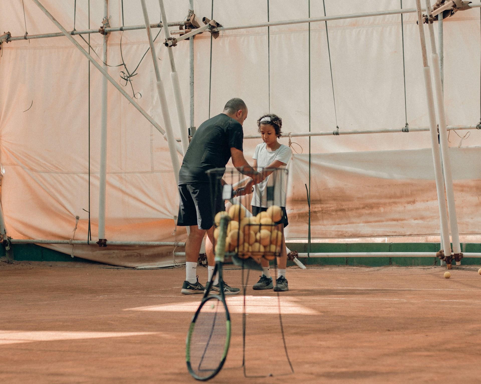 A tennis coach, wearing black, shows a pupil wearing a white tee with black shorts how to hold their racket, behind a basket half full of balls.