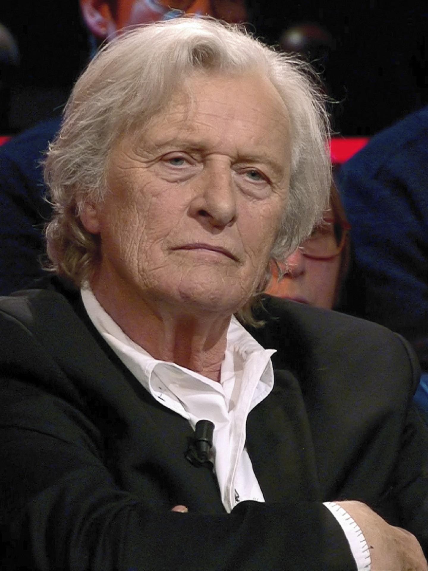 A man with blond-silver hair wears a black jacket over a white shirt.