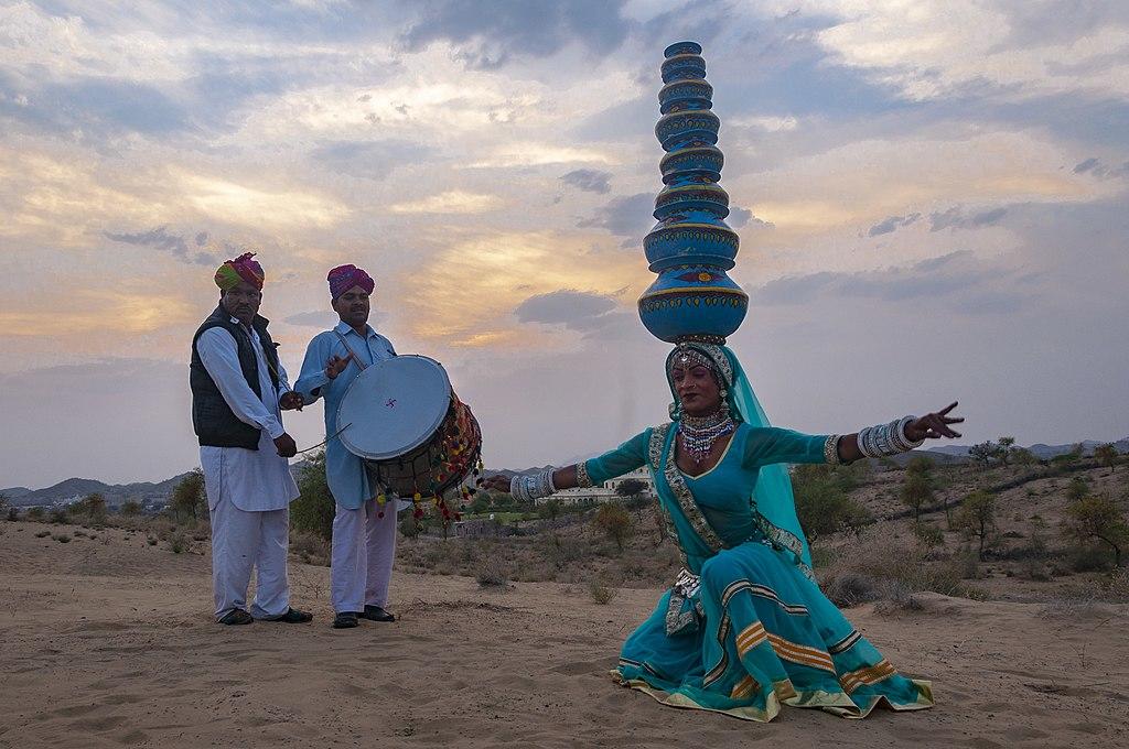 An Indian woman in traditional Saree balances a stack of earthen pots on her head while dancing. Two men play instruments in traditional garb.