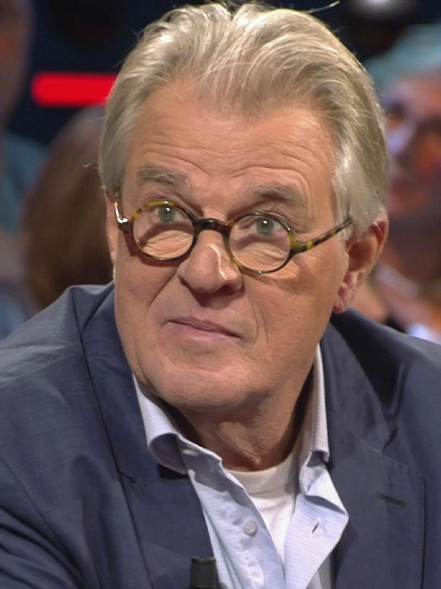 A blond man wears glasses and a charcoal grey jacket over a white shirt.