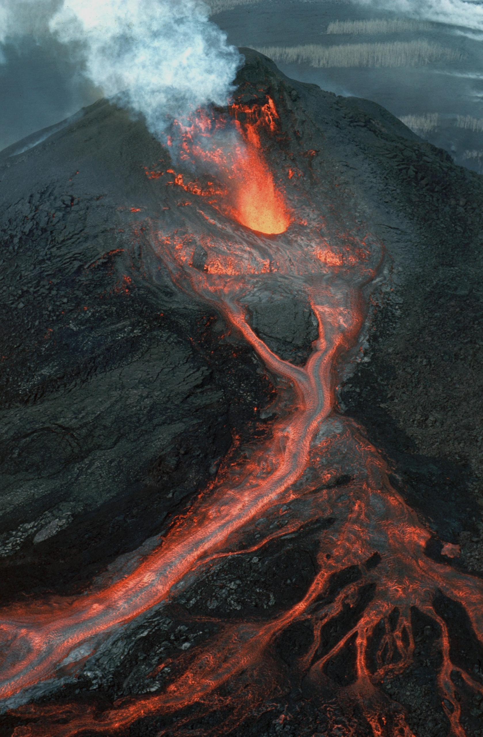 A volcanic eruption at dusk, with red lava flowing down the volcano's side and a plume of smoke above it. 