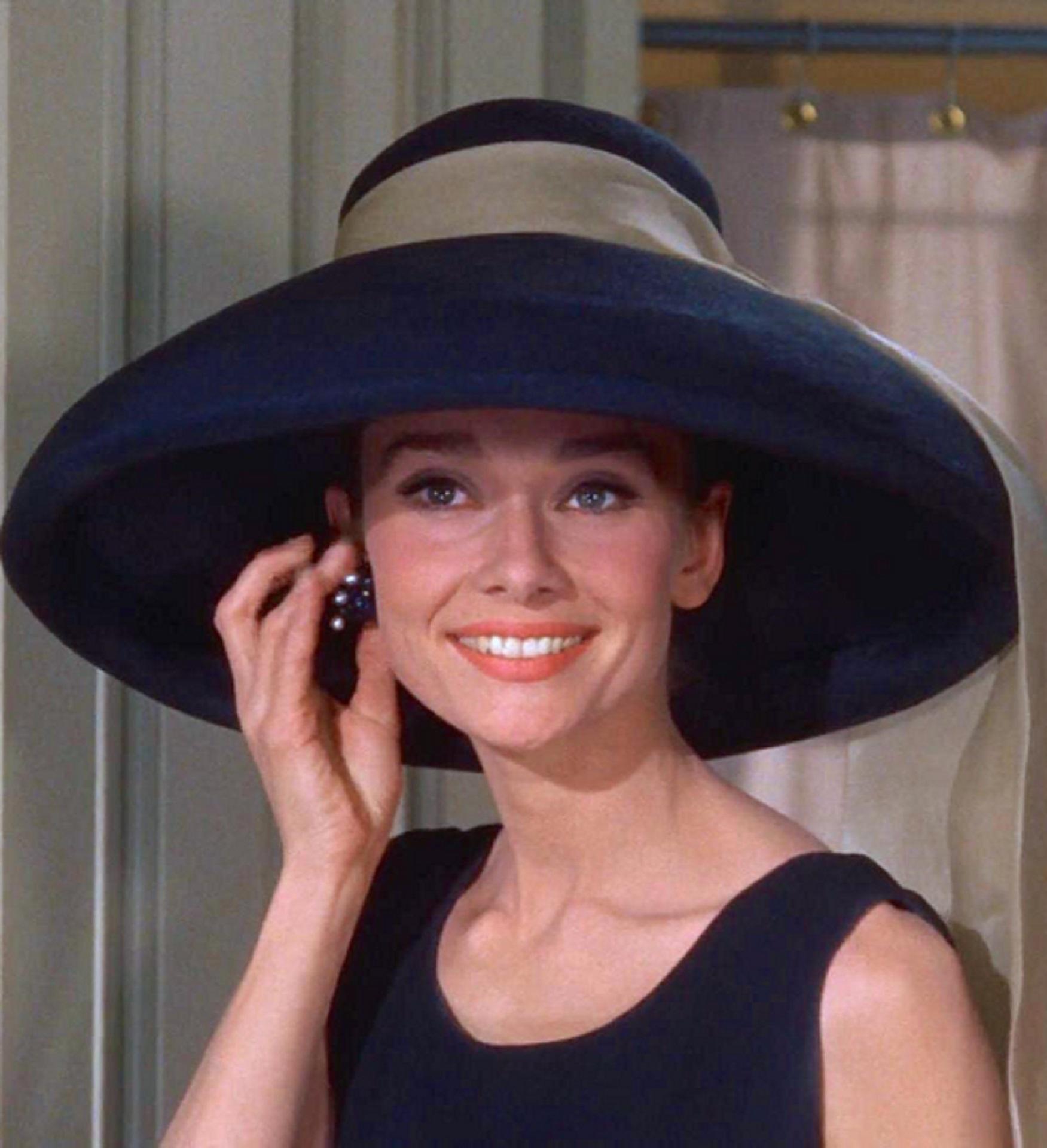 A woman wearing a black dress and a large black had holds her hand to her ear and smiles.