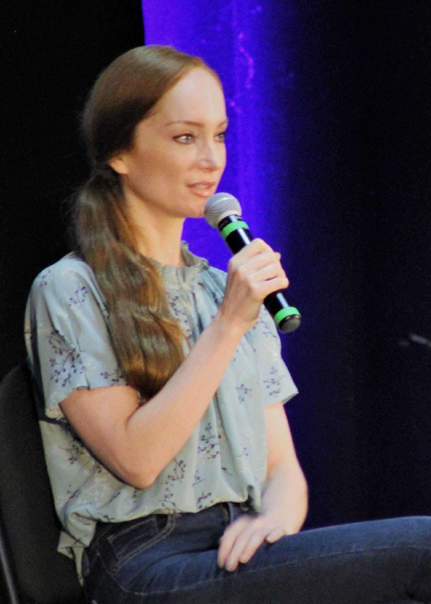 A woman wearing a green shirt and dark trousers holds a microphone. 