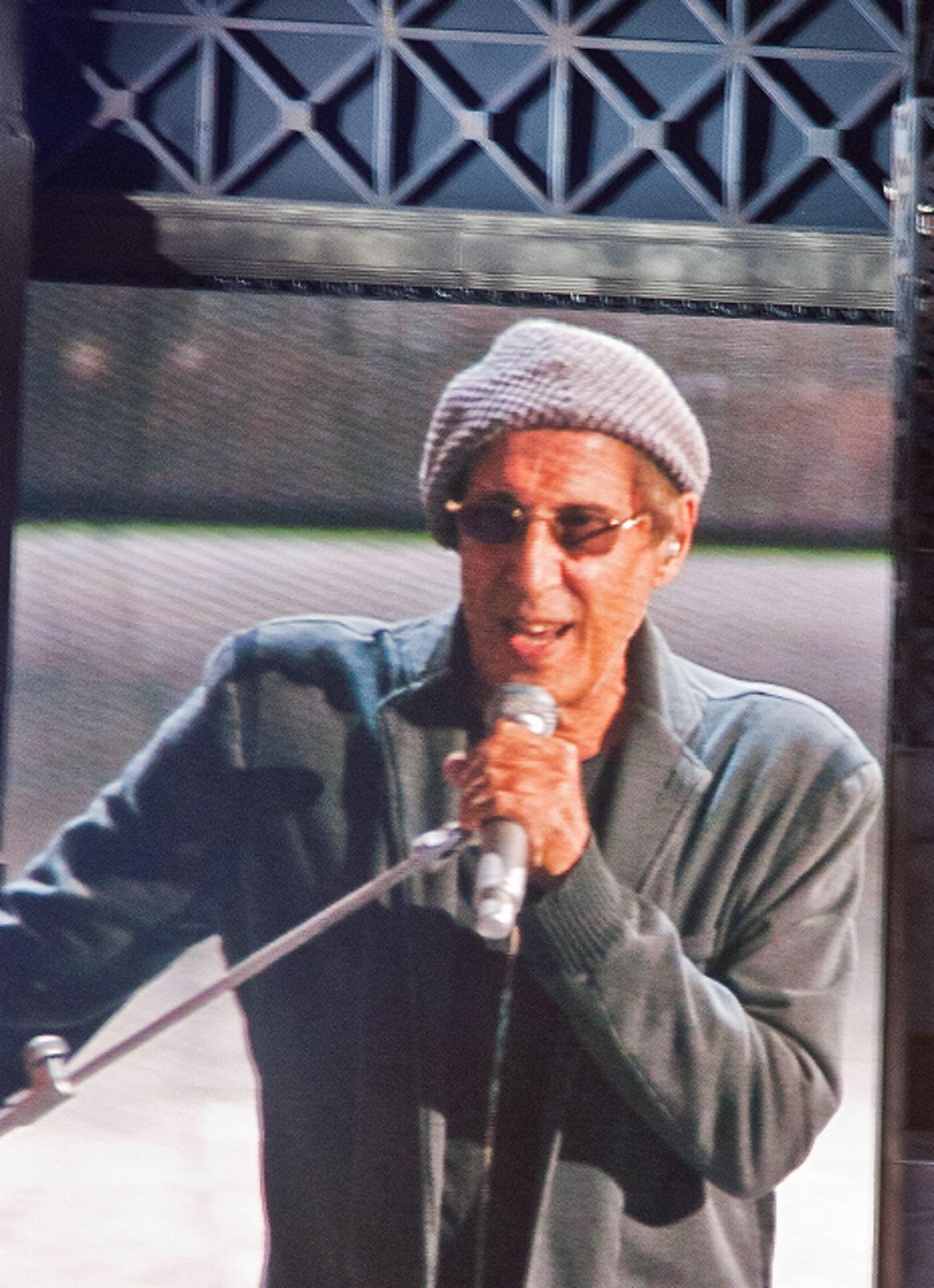 A person wearing a beret and a grey jacket holds a microphone.