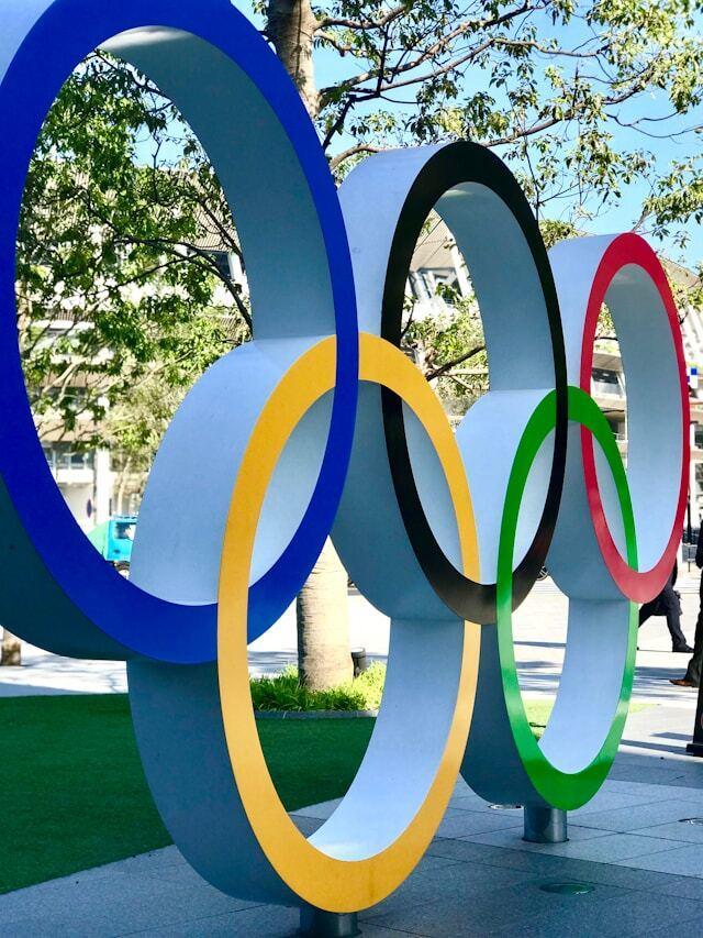 Outdoor sculpture of the Olympic symbol, displayed in an urban setting with trees and a sidewalk in the background.