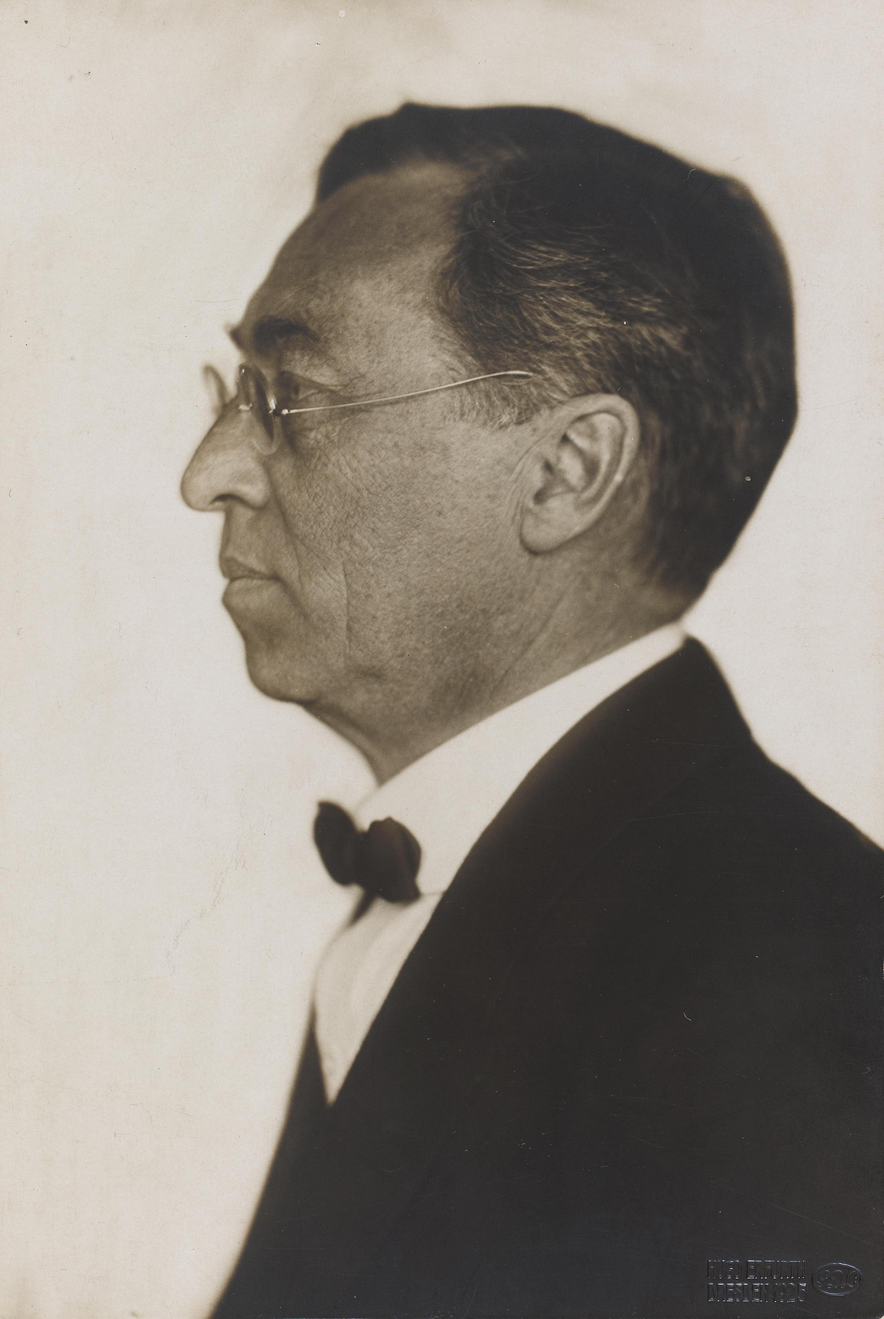 A man wearing a suit with bow tie and glasses, stands in profile.