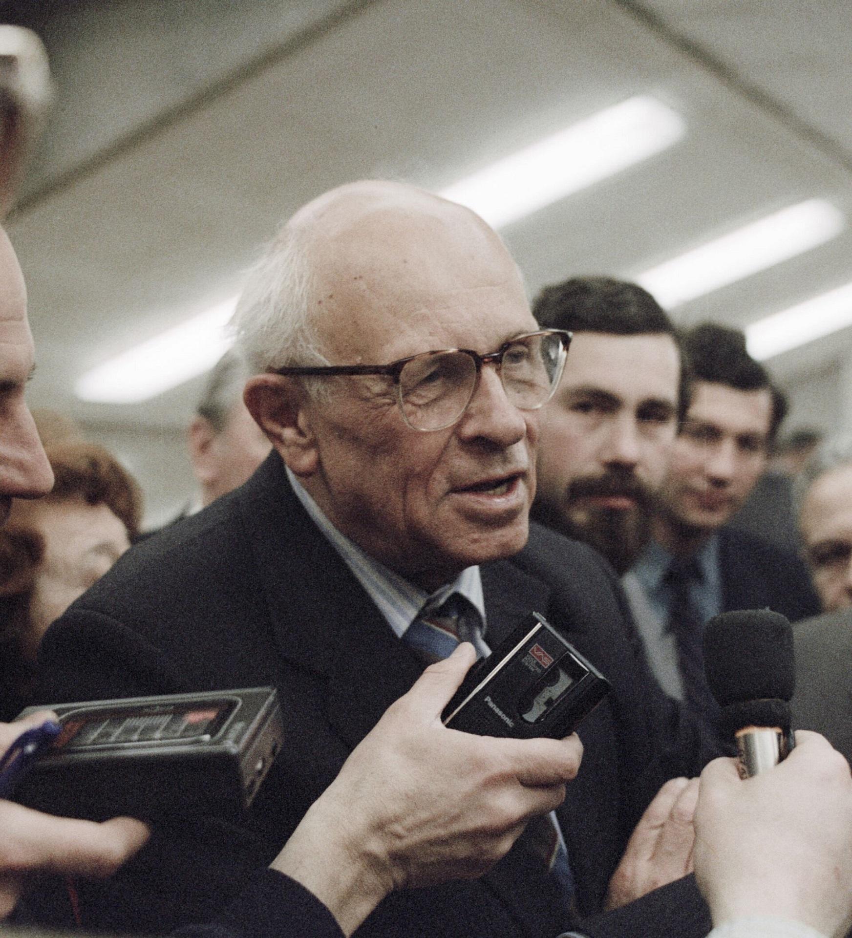 An elderly man wearing a suit and tie talks with people holding microphones.