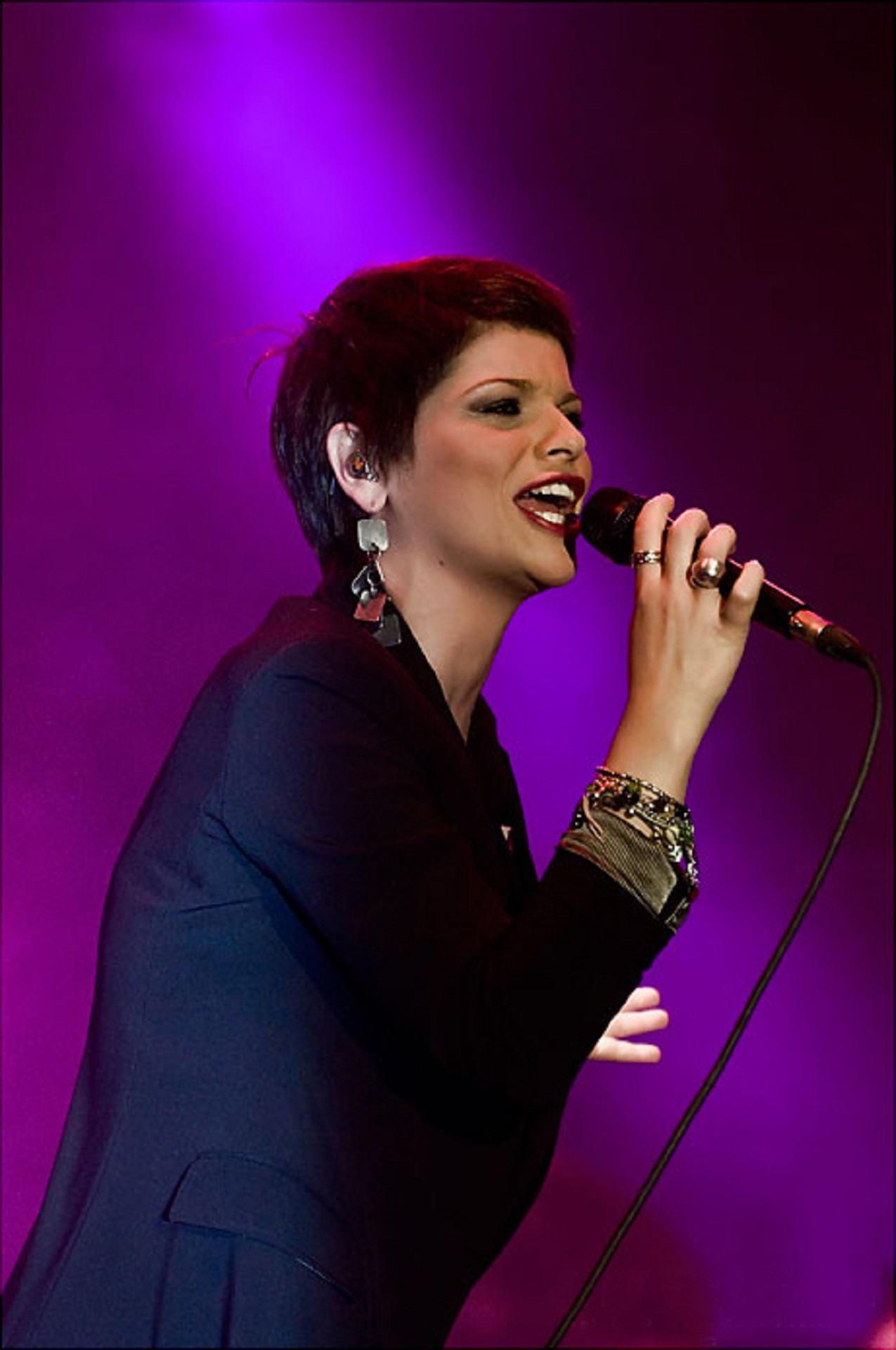 A woman wearing a black jacket holds a microphone on stage, against a purple background.