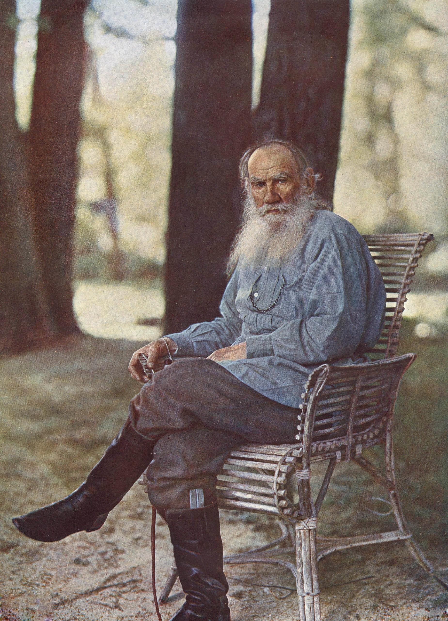 A bearded man in a blue shirt and grey breeches sits on a wooden chair, surrounded by trees.