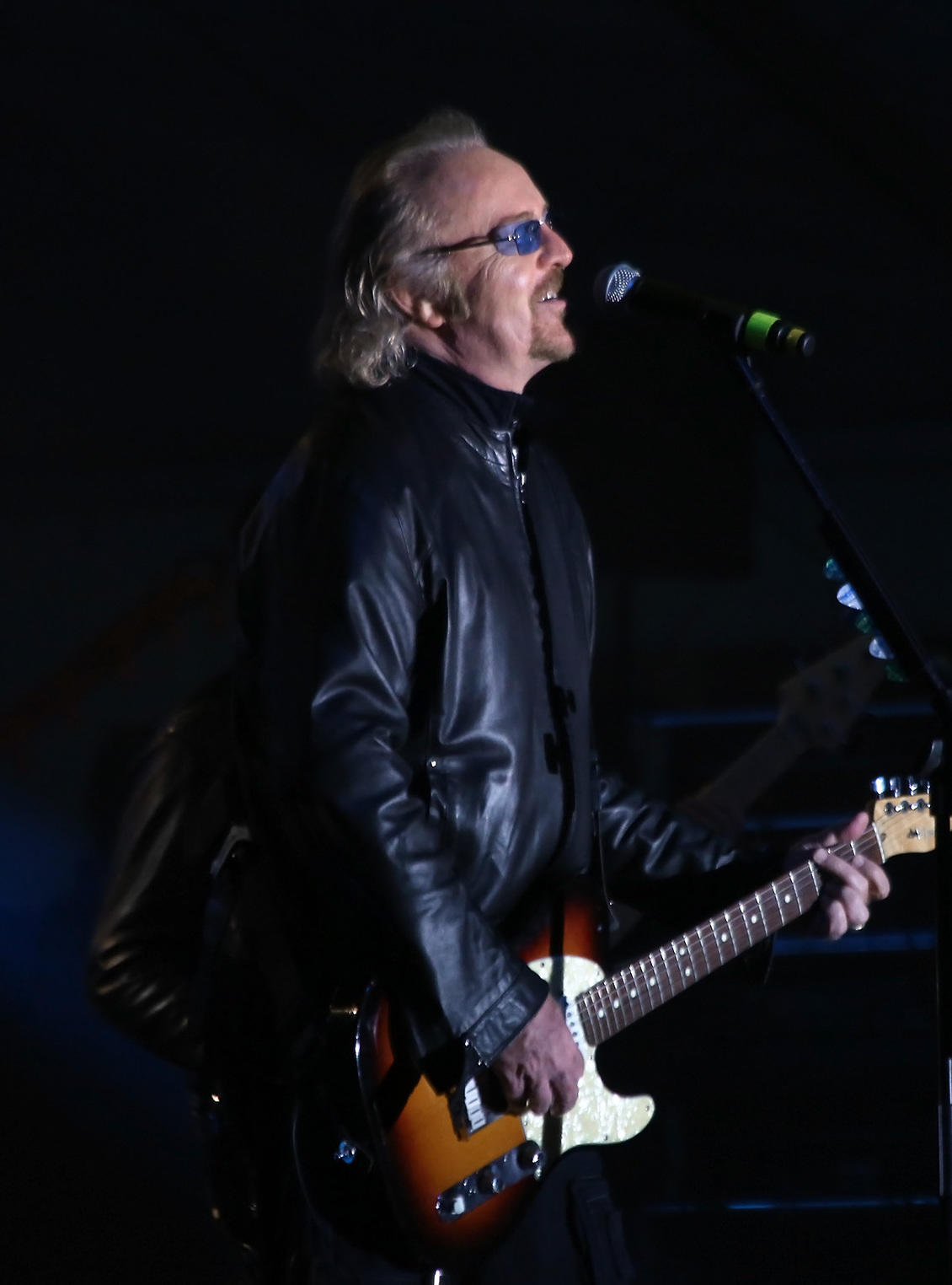 A man wearing sunglasses and a leather jacket holds a guitar and smiles.
