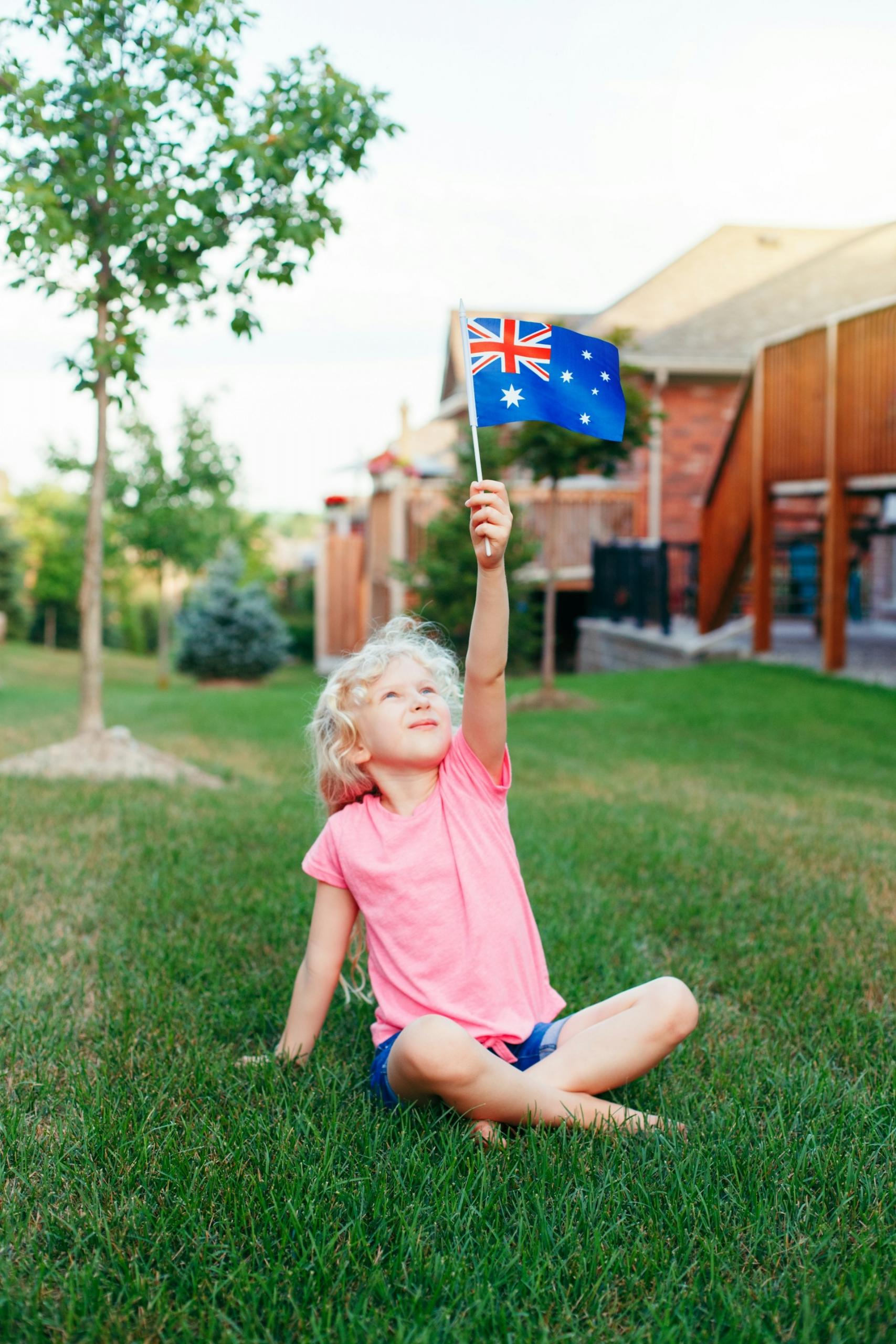 A small child holds up an Australian flag while sitting on a green lawn. 