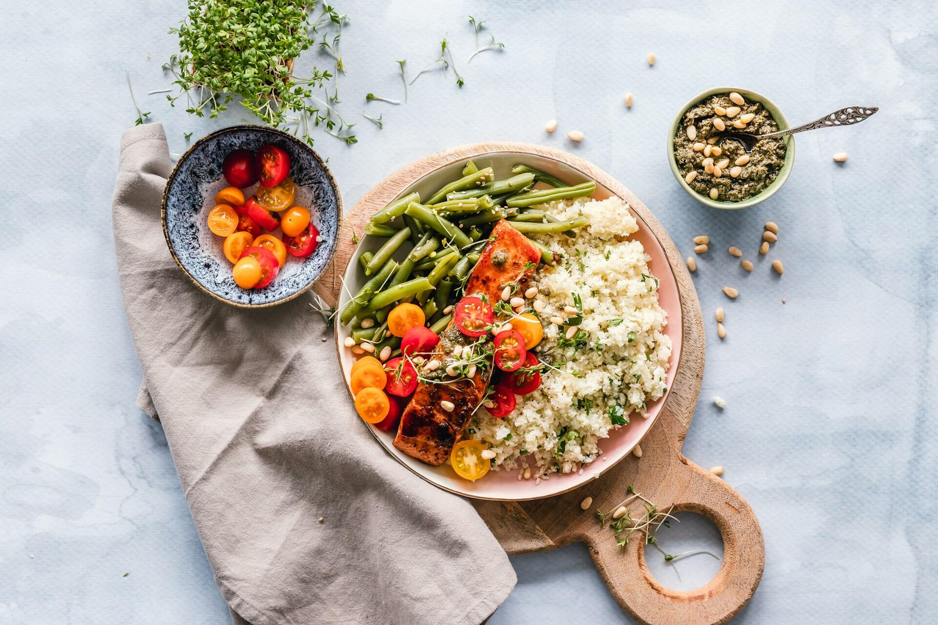 healthy meal of rice, salmon, tomatoes, and green beans with more tomatoes, homemade pesto, and sprouts spread around the plate, which rests on a wooden paddle on a white background
