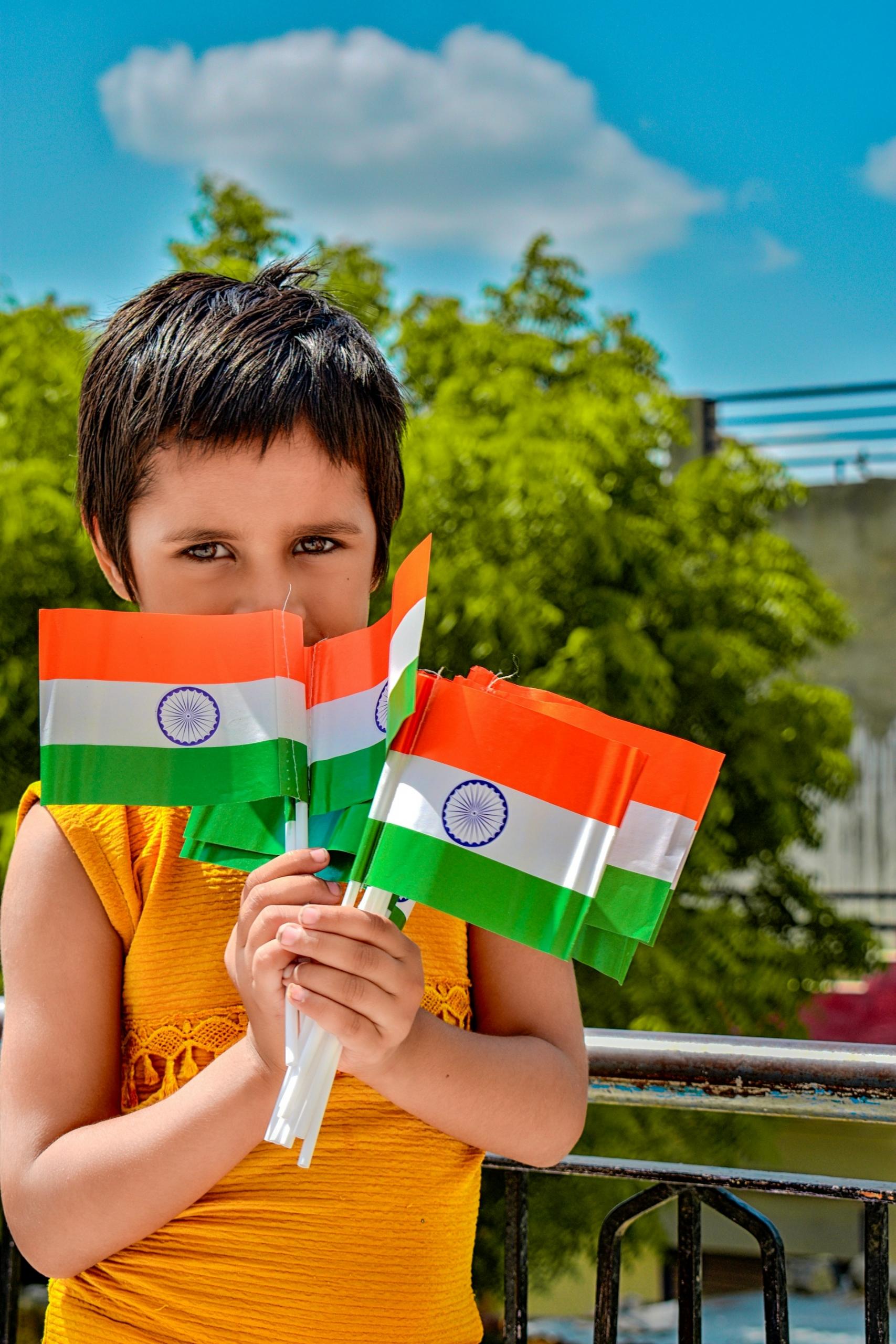 A boy in an orange shirt holds Indian flags on a sunny day.