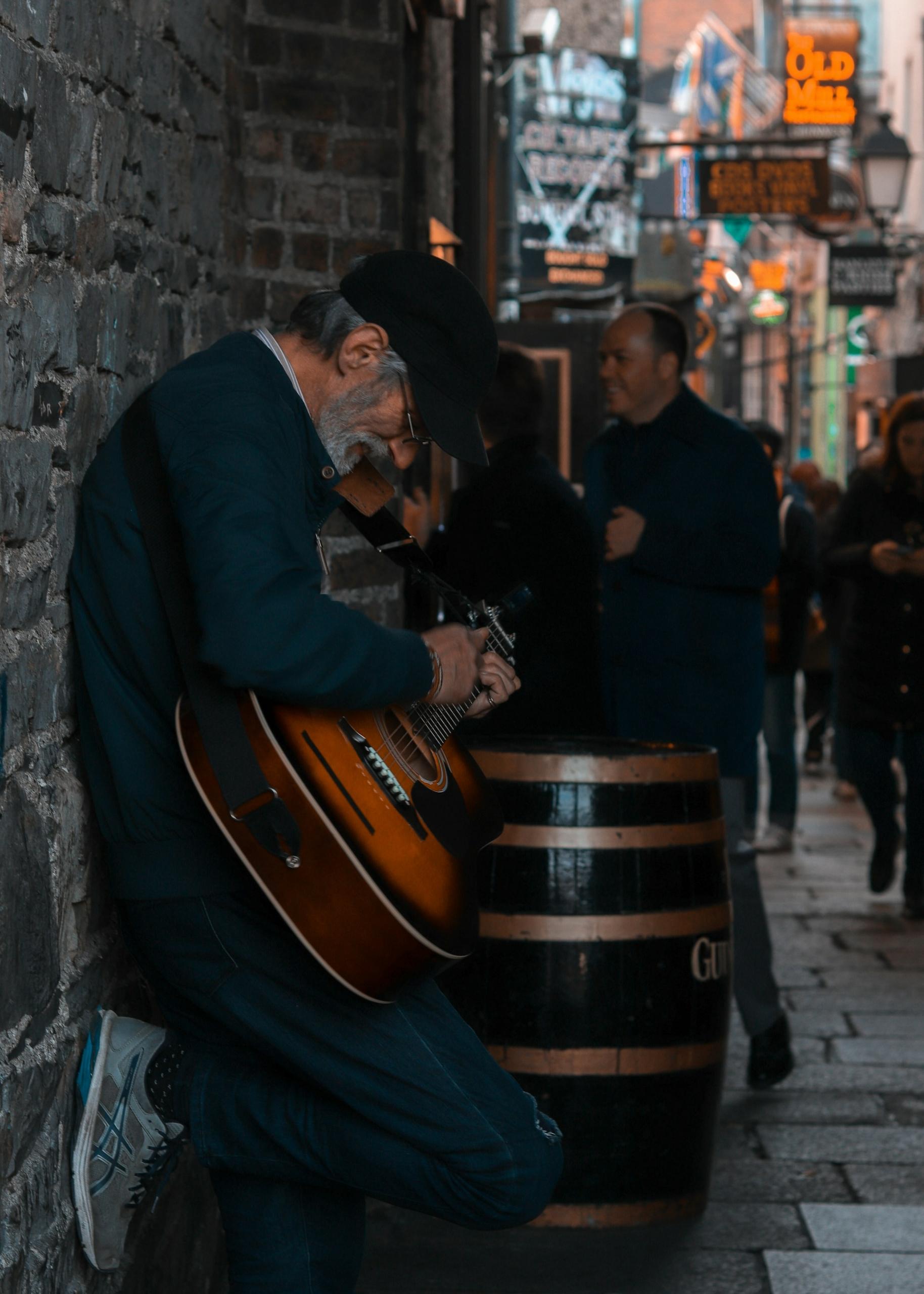 A man holds a guitar while leaning against a wall in a dark alley during the day.