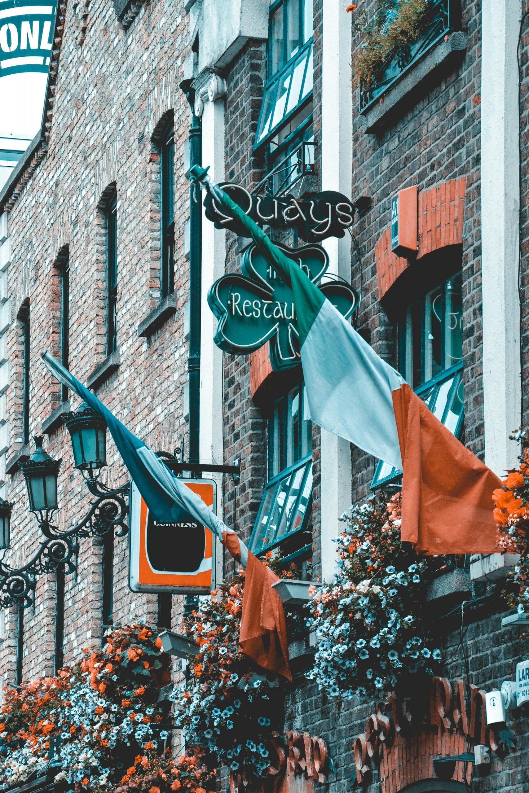 Two Irish flags mounted on a stome building next to a neon shamrock sign. 